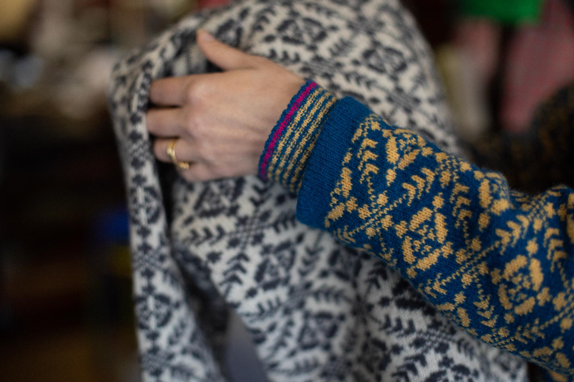 A studio assistant at Nielanell Shetland knitwear helps a customer try on a Fair Isle Style Shetland jumper. Just hands are seen. The assistant wears a yellow and blue patterned jumper, the one being tried on is an off white  Fair Isle Jumper with dark grey patterning.