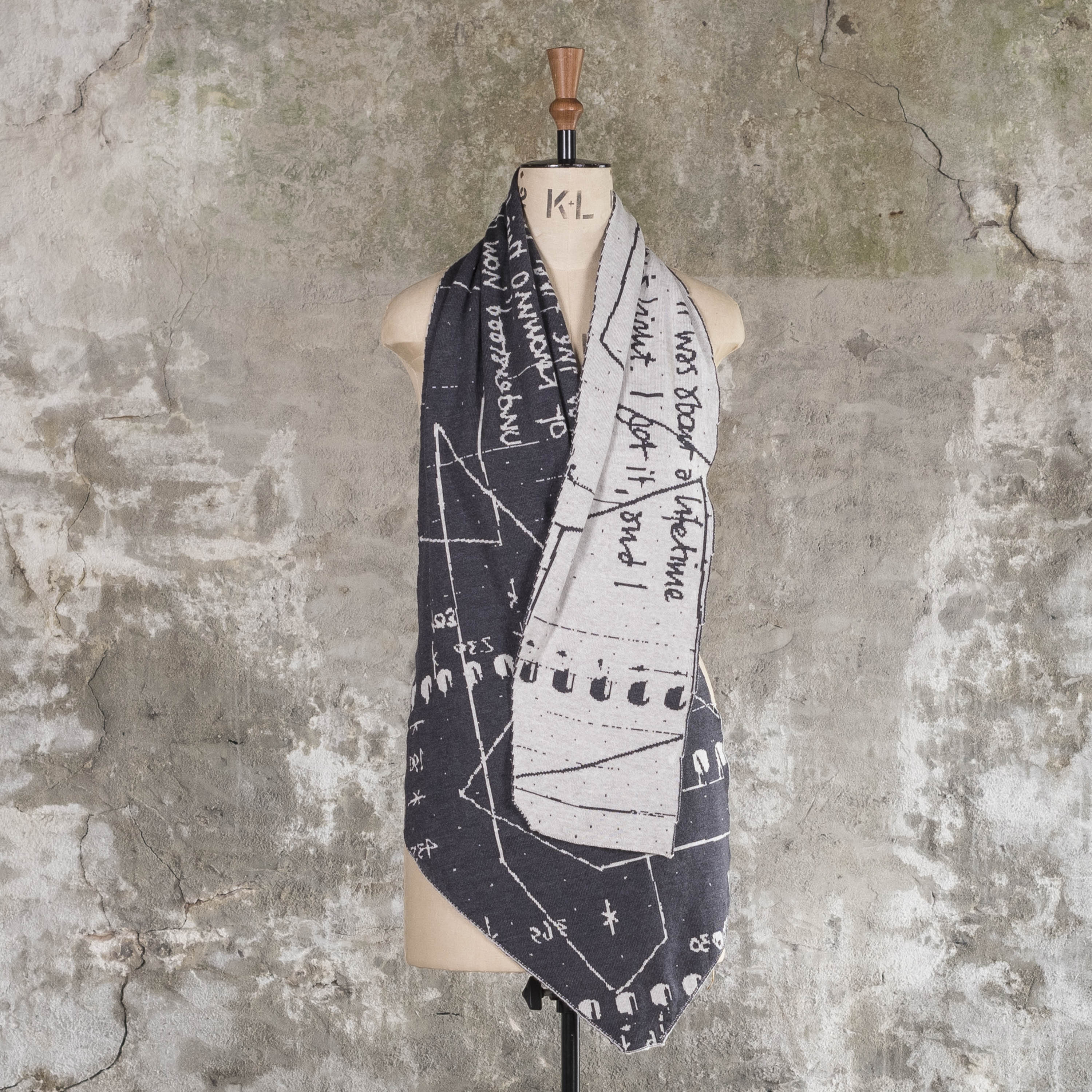 Contemporary, irregular shaped scarf with abstract pattern, knitted in charcoal grey and stone white. Shown on vintage mannequin against Shetland rustic wall.