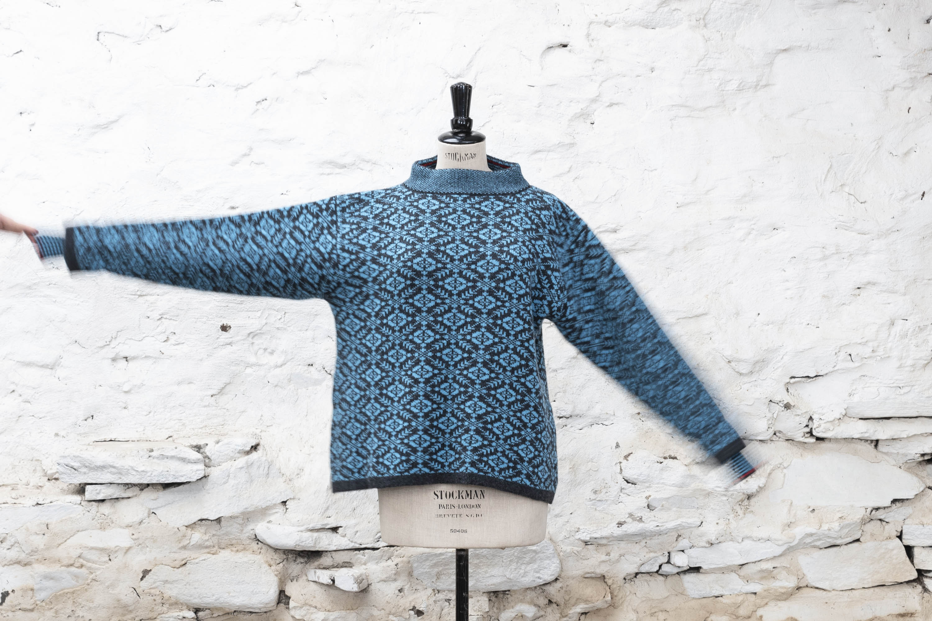 Real Shetland-made Fair Isle jumper in a bright turquoise and navy. Front view against a white-washed wall, with the right arm held up. The pattern is nordic-inspired all-over geometric pattern.