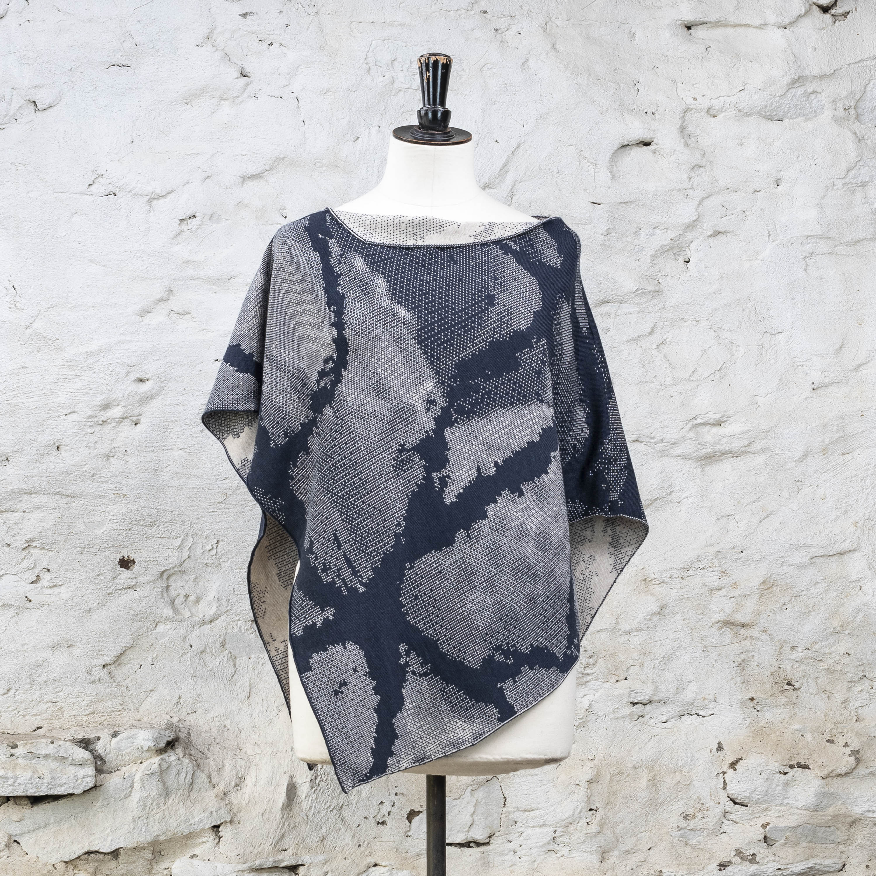 Rani knitted cape in abstract pattern in inky blue and antique white. Shown on a vintage mannequin against a whitewashed rustic stone wall