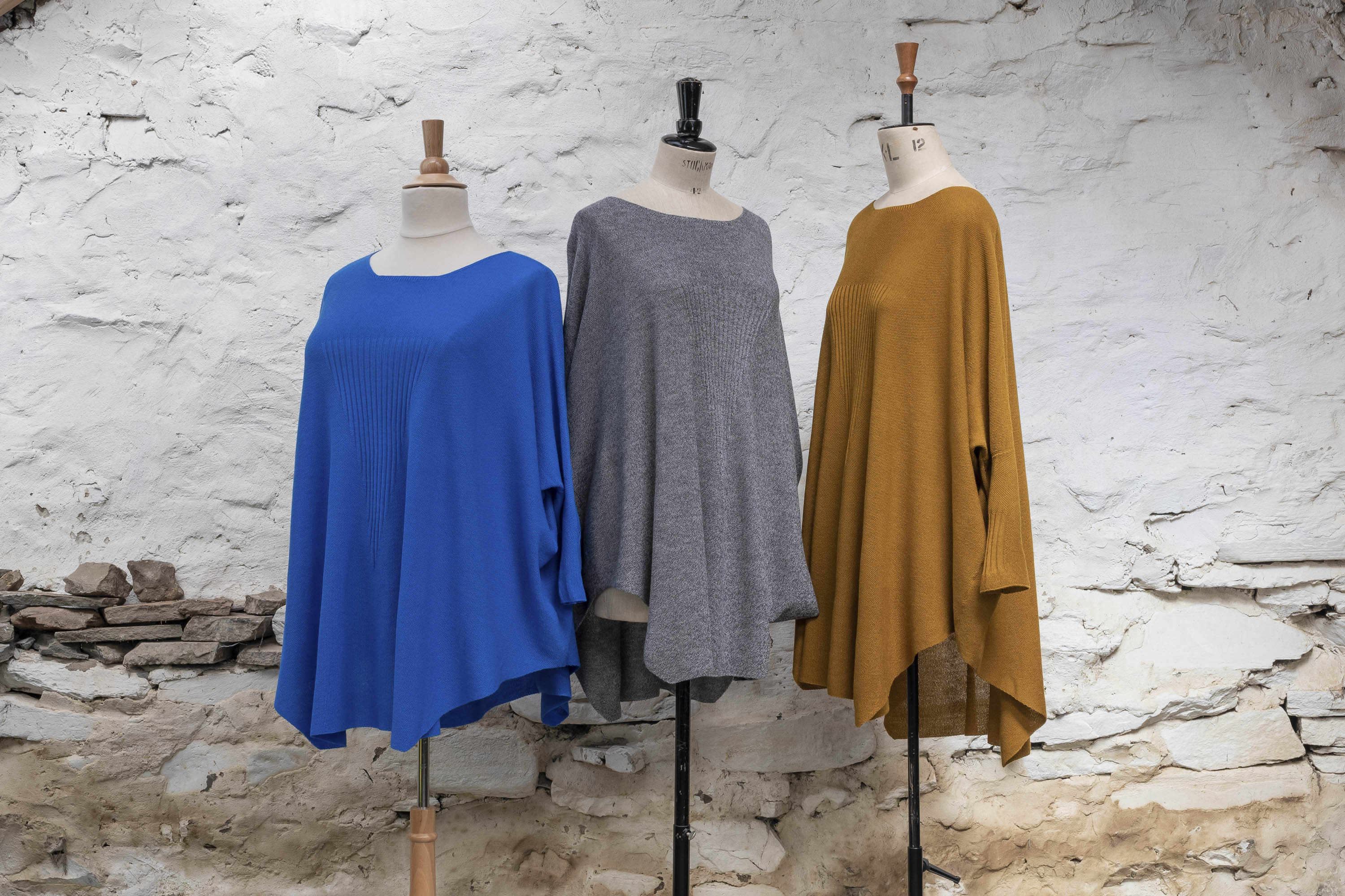 Three easy, loose and lightweight summer jumpers on vintage mannequins shown against a whitewashed rustic stone wall. One is in a deep electric blue, one in a marled mid grey and another in yellow ochre.