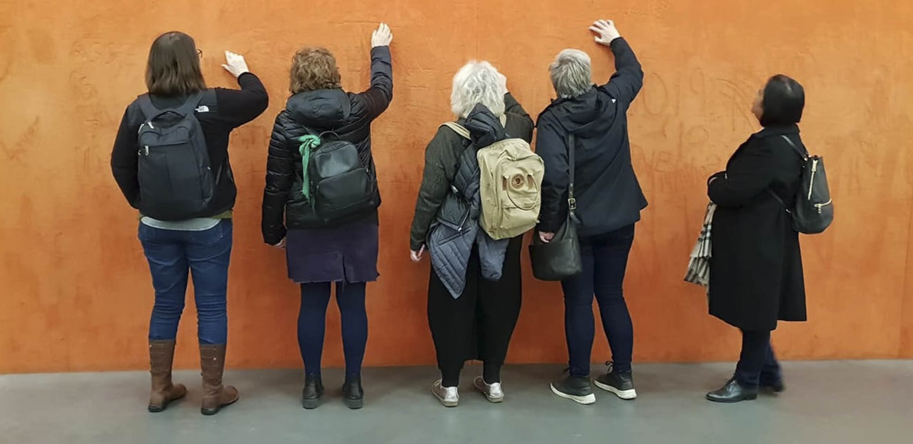 Five women stand with their backs to the camera, drawing with their fingers on a wall covered with orange pigment