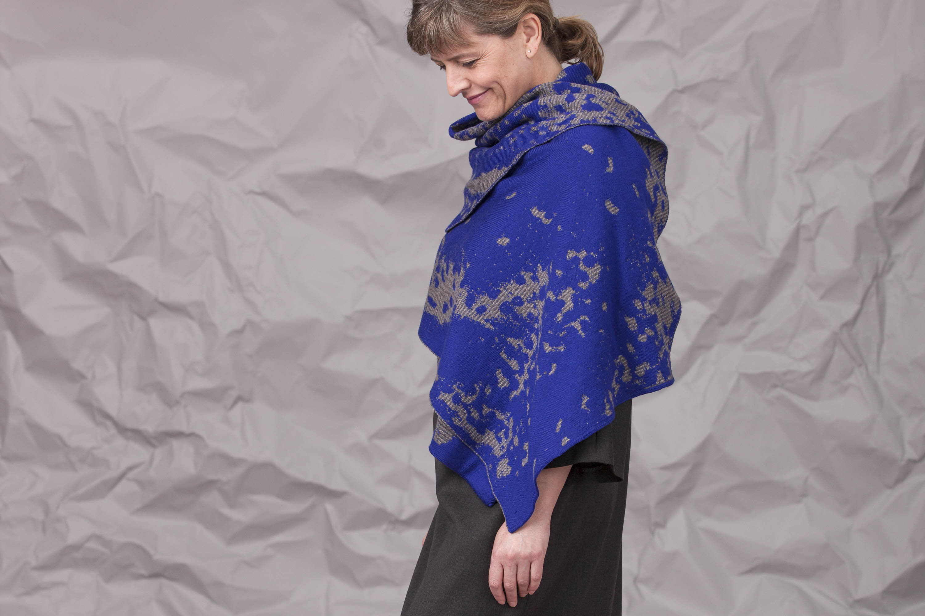Modern Shetland shawl worn over a charcoal grey fine wool dress. Model stands side on with abstract knitted wrap in Yves Klein blue and greys
