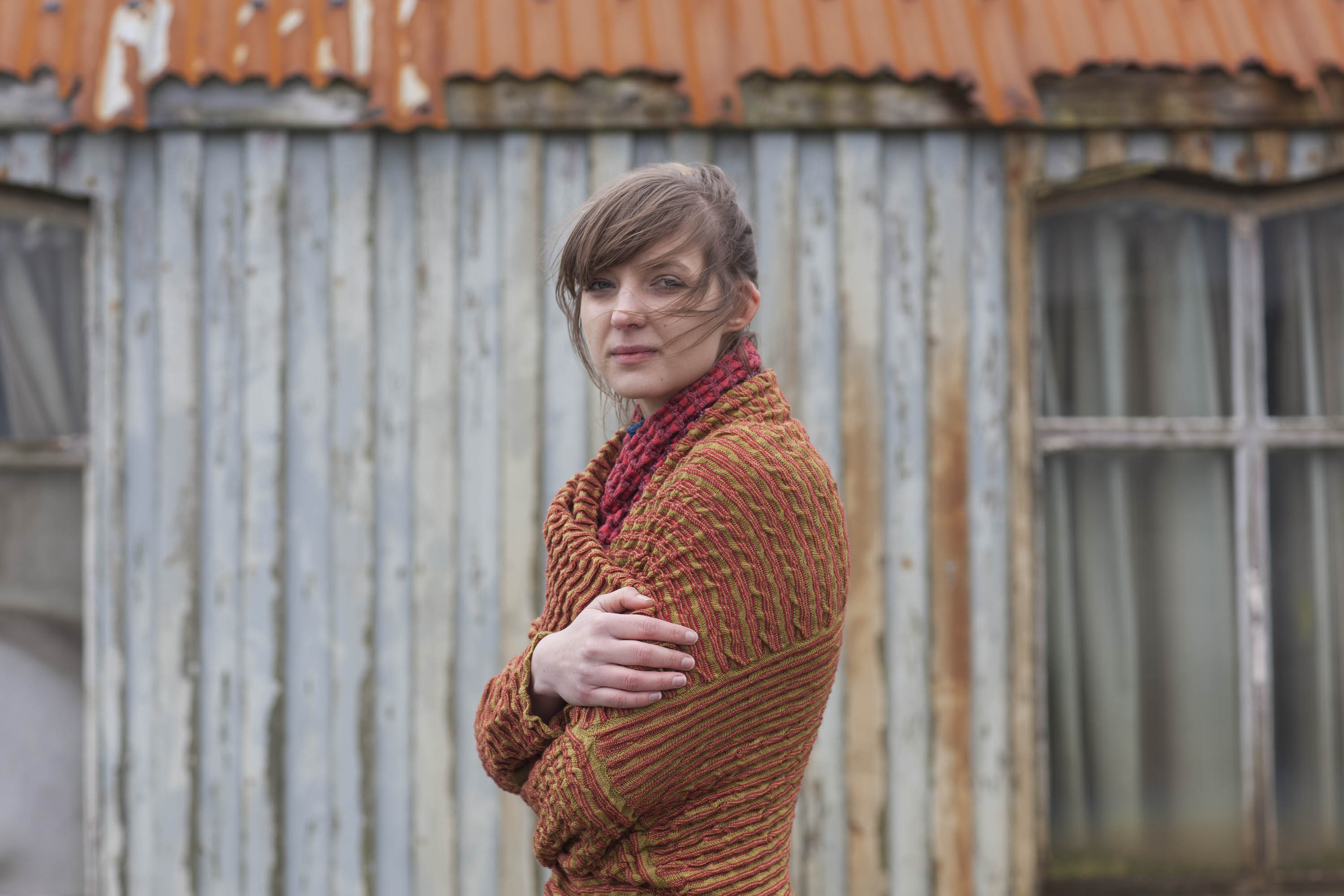 Contemporary Shetland knitwear in Hoswick, a model wears a rusty, textured knitted jacket with her arms wrapped round herself and her hair being blown in the wind