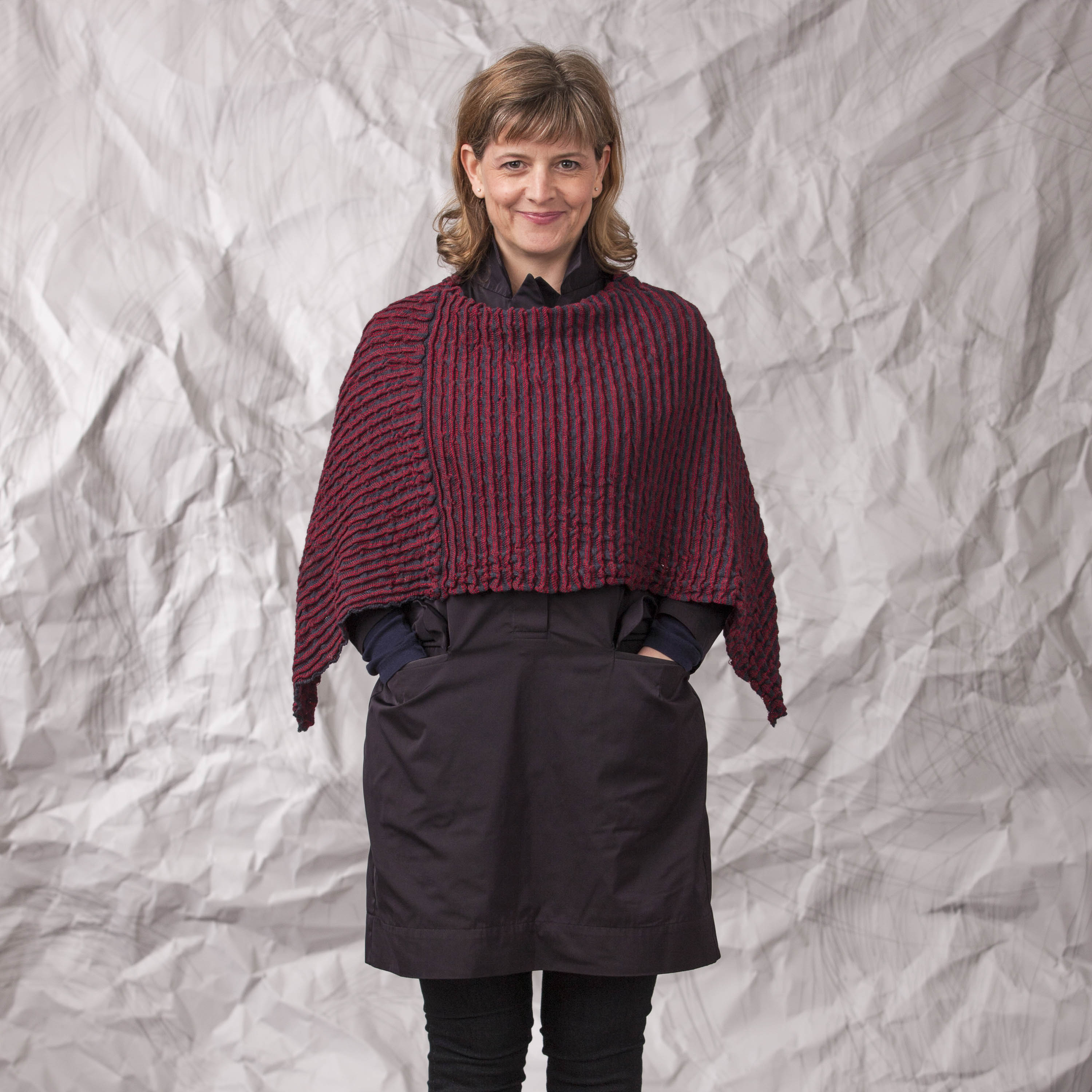 Model wears a lightweight contemporary poncho made in Scotland. It has lightly ridged texture knitted in cherry and navy.