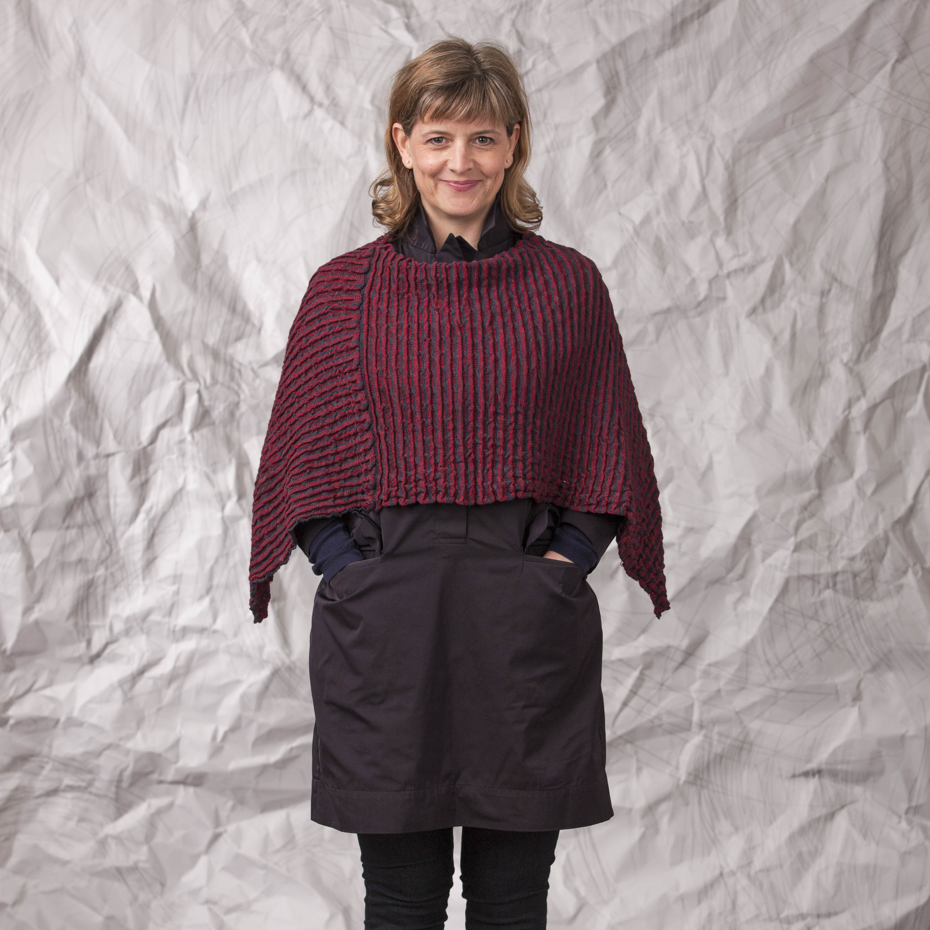 Contemporary Scottish knitwear, poncho in gently ridged fabric in dark berry coloured. Worn by a woman, over a navy shirt dress