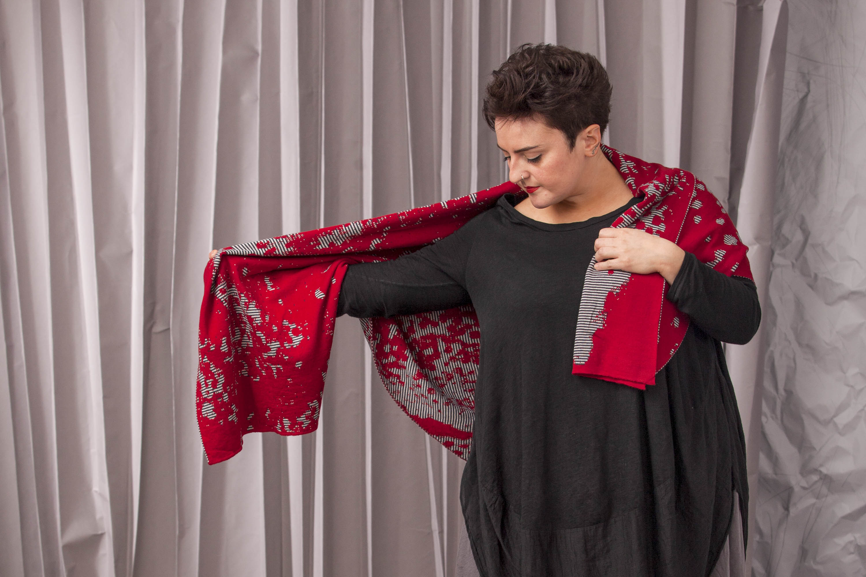 A woman in a loose, black dress wears a large abstract patterned modern Shetland shawl in reds and greys