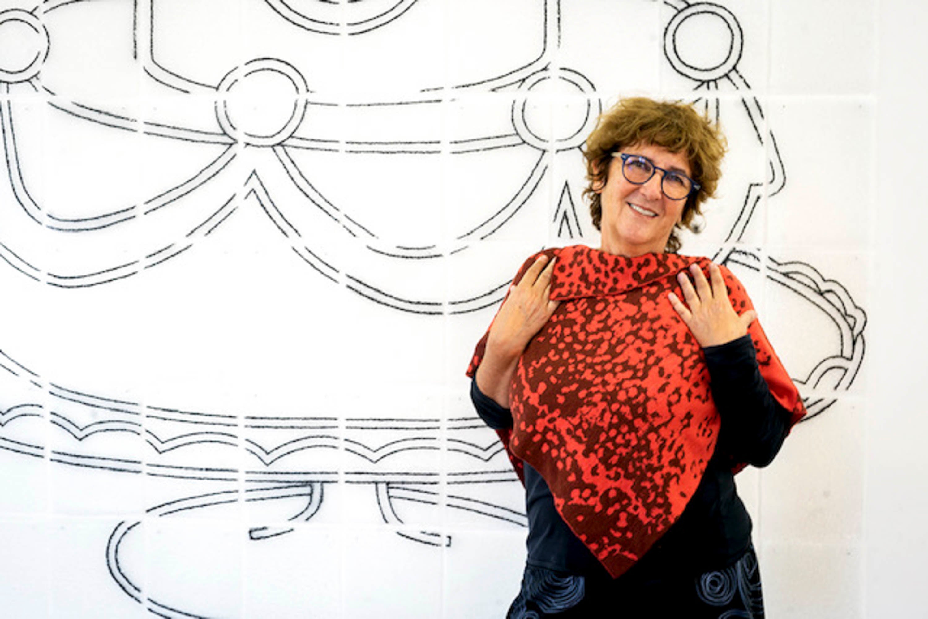 A woman stands against an abstract mural, wearing a cape in two rust colours. She holds her arms up to her collar bone