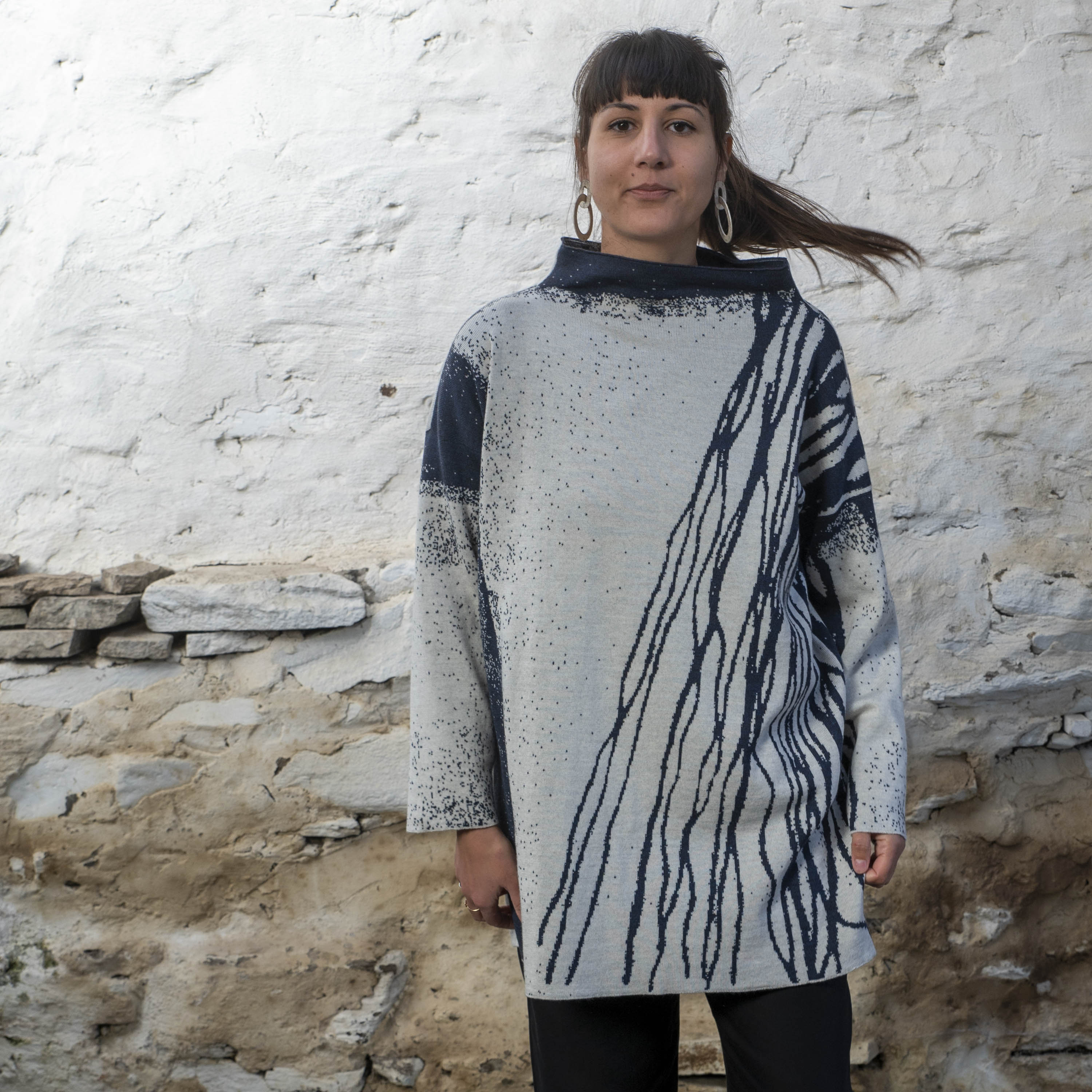 In a stone built, rustic Shetland building with whitewashed walls a woman with long dark hair tied back wears a longline tunic jumper in an abstract linear pattern. It is off white and navy. She wears black trousers and swings here hair so that her pony tail flies out