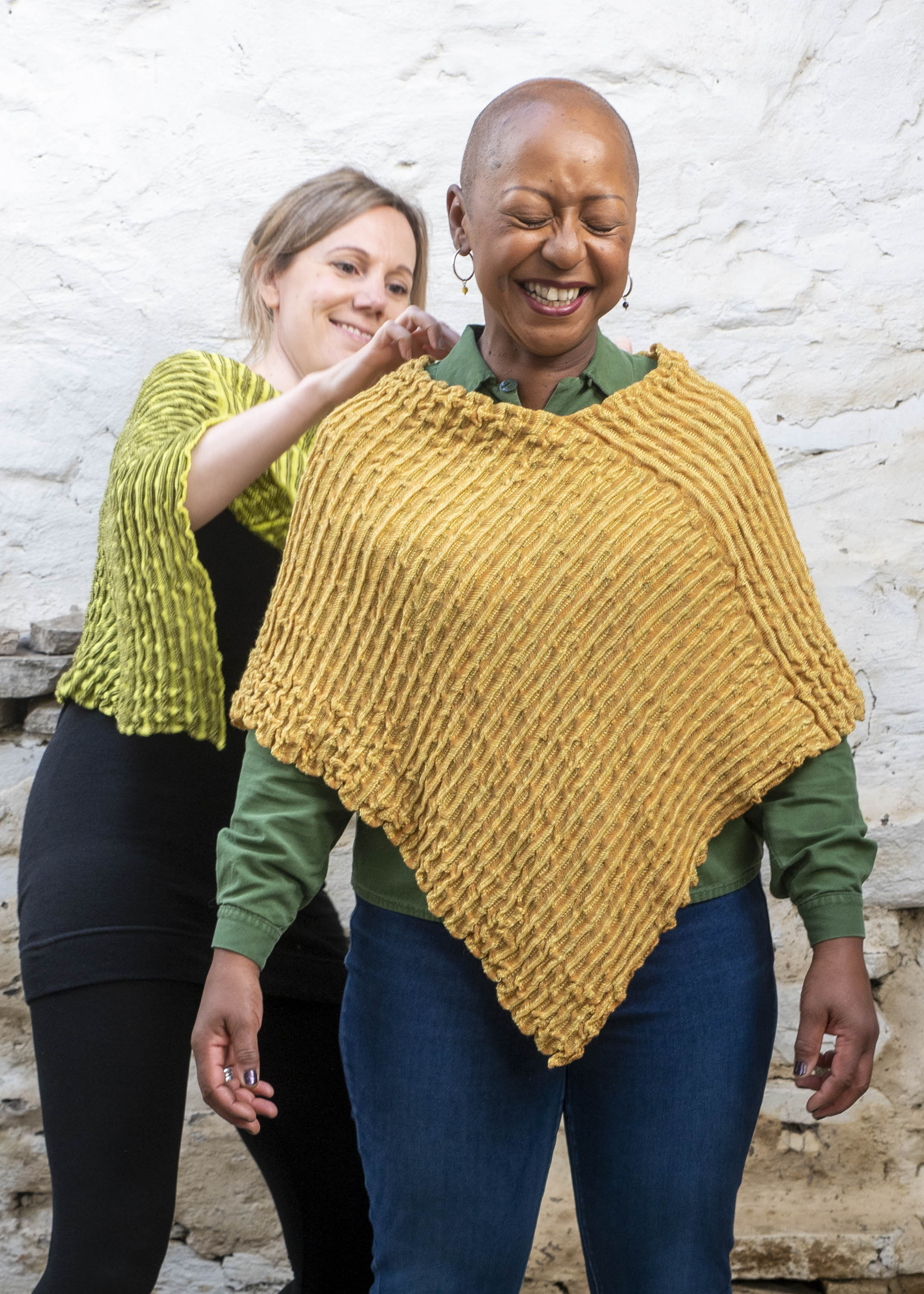 Two women wearing softly ridged, knitted capes. In the foreground, a black woman wears a golden cape over a green shirt and jeans. In the background a white woman with hair tied back wears a green and yellow cape. She adjusts the woman in front's cape. They are both smiling and laughing. Photograph taken inside a rustic Shetland stone shed, whitewashed.