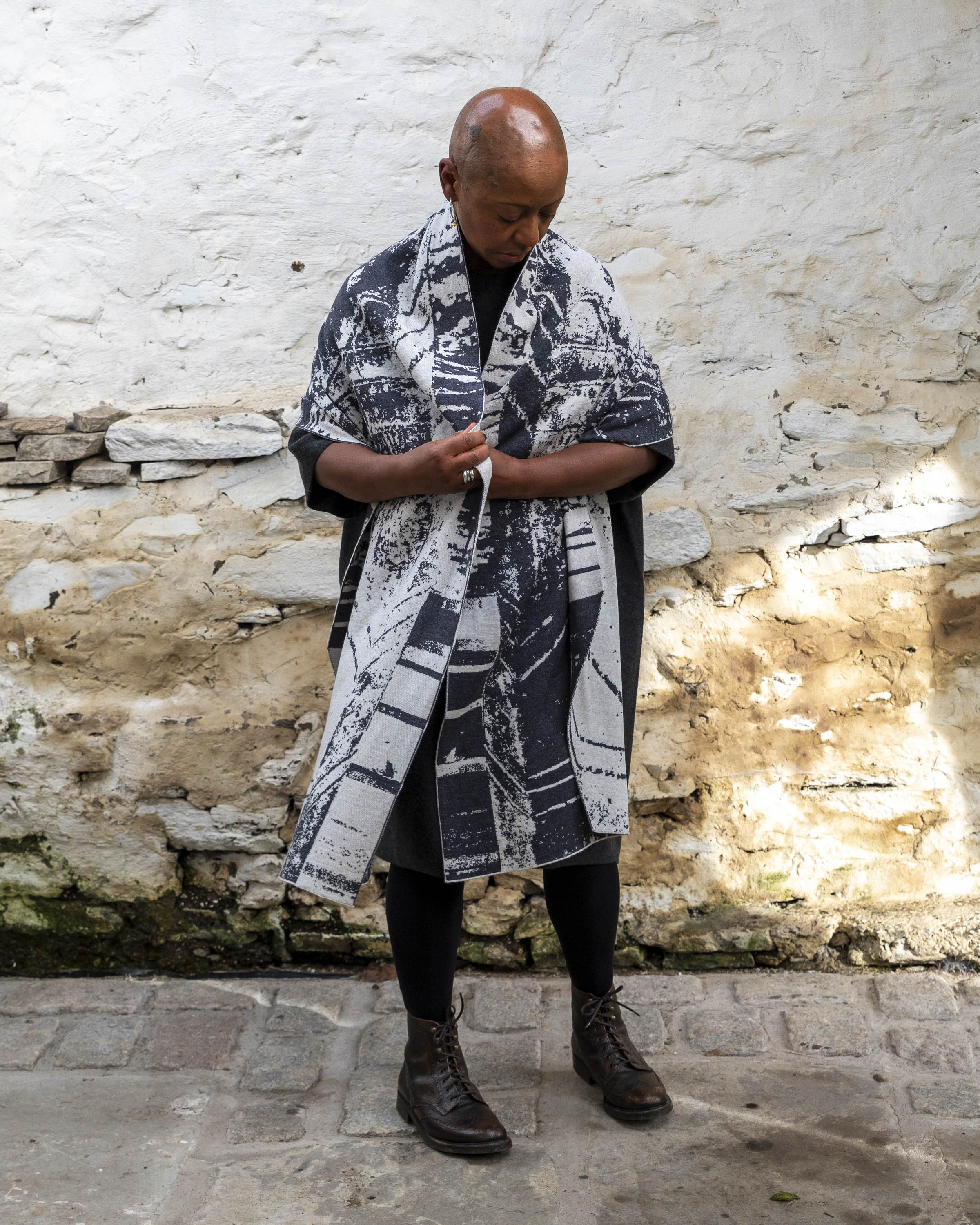 A black woman with a shaved head stands in a rustic stone building with whitewashed walls in Hoswick, Shetland. She wears black tights, a black charcoal woollen dress and wraps a large off white and charcoal shawl around her shoulders and draped down, almost looking like a coat or cape.