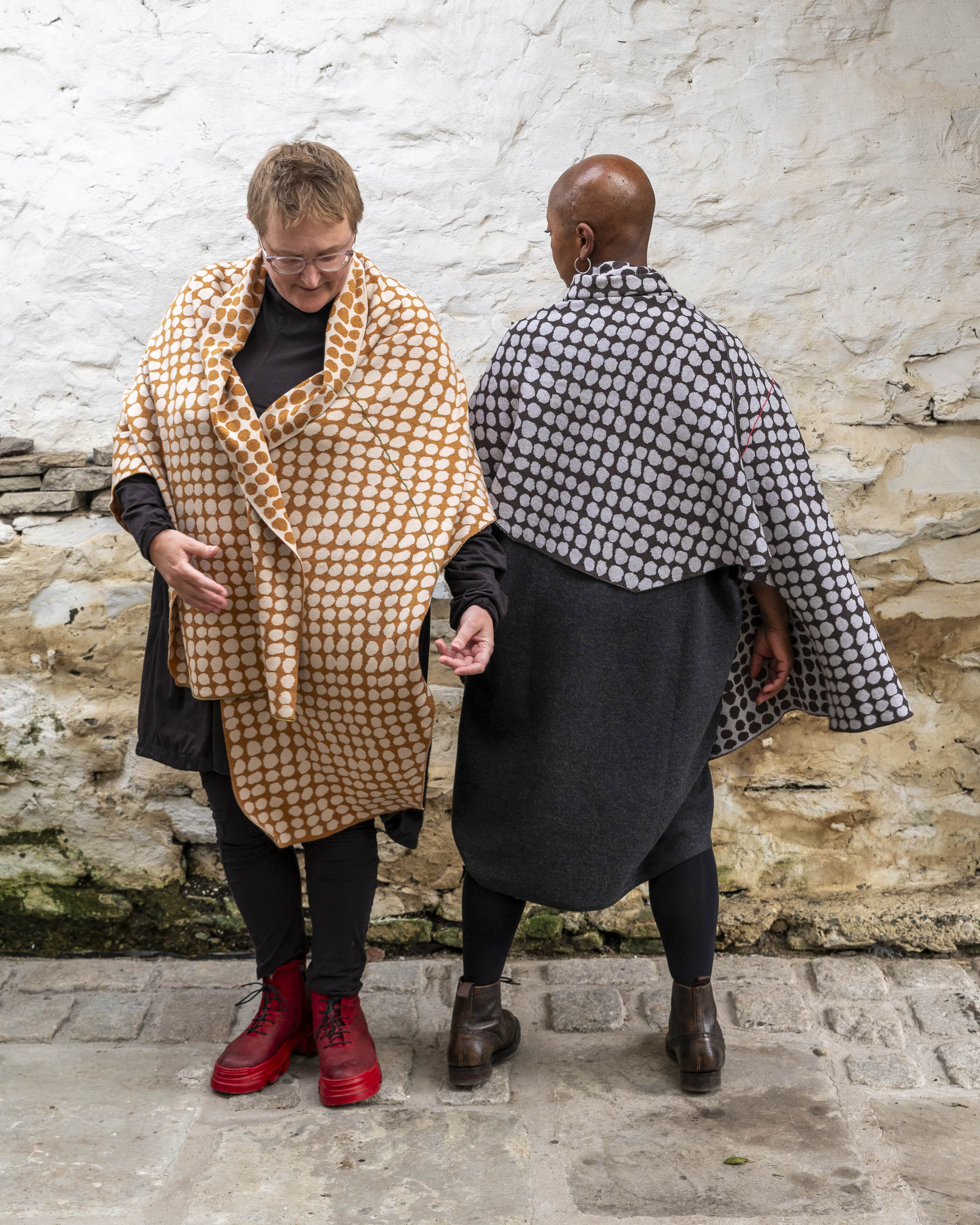 Two women stands in a whitewashed stone shed with flagstoned floor. On the left a white woman with fair hair wears a modern shawl in an ochre colour with irregular dots over a black tunic and leggings. On the right, a black woman with a shaved head wears the same shawl but in charcoal grey and pale grey over a grey woollen dress.