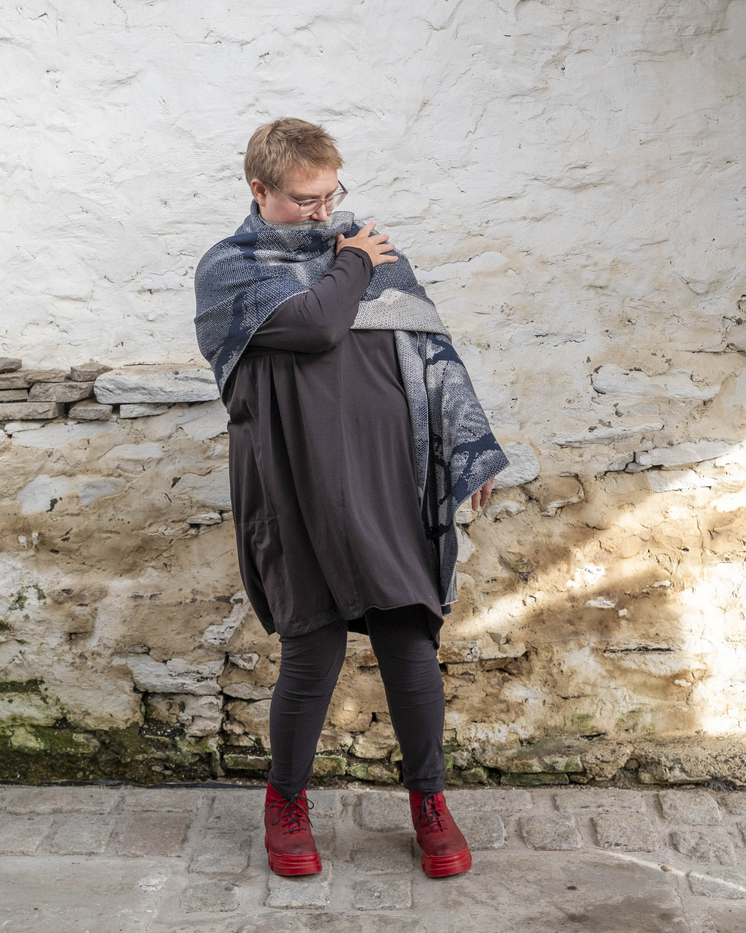 A white woman with short, fair hair stands inside a rustic stone building in Hoswick, Shetland. She wears an off-black tunic with matching leggings and red platform boots. She wears a navy and off white oversized shawl, she has the shawl around her shoulders and holds one arm up to her shoulders.