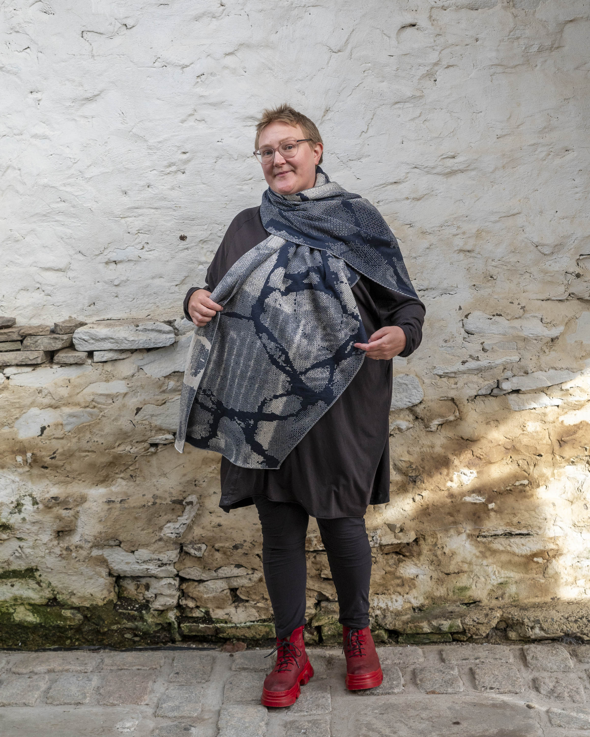 A white woman with short, fair hair stands inside a rustic stone building in Hoswick, Shetland. She wears an off-black tunic with matching leggings and red platform boots. She wears a navy and off white oversized shawl  which is arranged like a giant scarf, and holds of the front to show the pattern