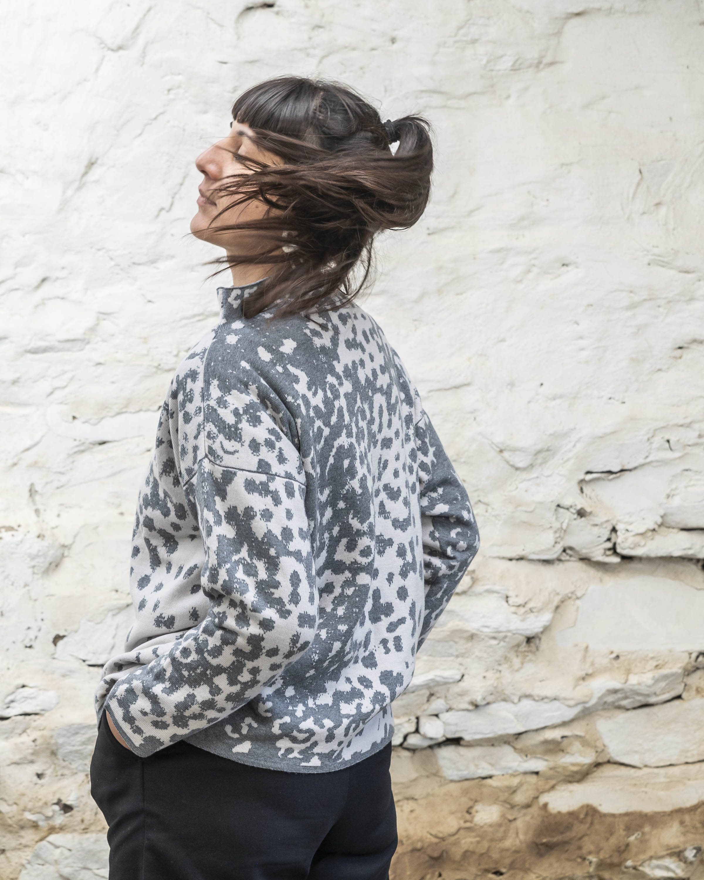 A woman with olive skin and dark hair tied back wears a charcoal grey and white mottled jumper. She is standing in a rustic stone shed with whitewashed walls and a flagstone floor. She is also wearing navy trousers and turns her head shaking her pony tail.