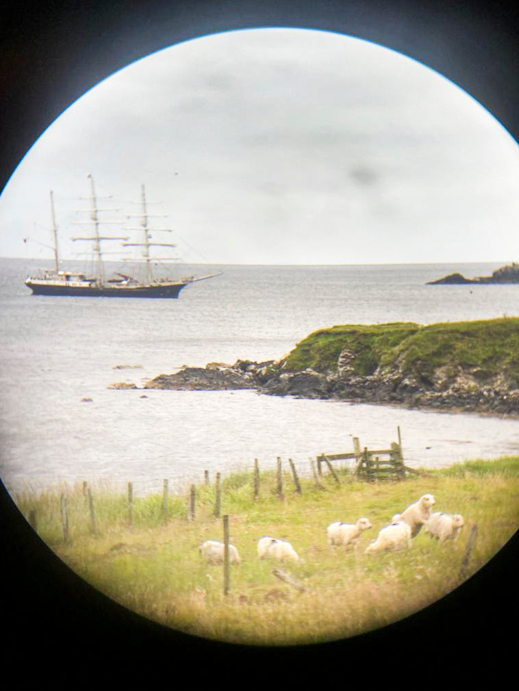 A photograph taken through binoculars of a tall ship with 3 masts  - its sails unfurled. The boat is offshore at Hoswick, Shetland with the shoreline and fields in the foreground.