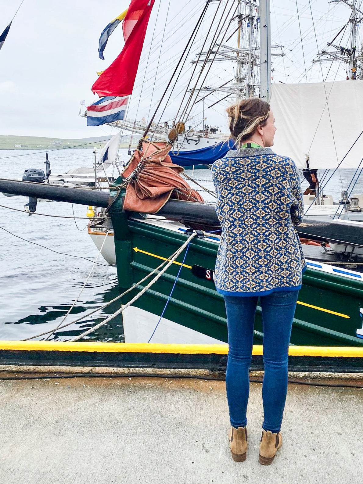 A white woman with her light brown hair tied back stands on the quayside in Lerwick. In front of her is Swan, a hundred year old timber boat. She is wearing a fair isle style jumper in an all-over pattern in blue and yellow, jeans and tan suede dealer boots.