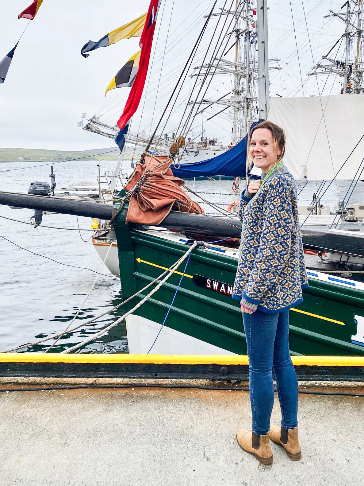 A white woman with her light brown hair tied back stands on the quayside in Lerwick. She turns and faces the camera, smiling. In front of her is Swan, a hundred year old timber boat. She is wearing a fair isle style jumper in an all-over pattern in blue and yellow, jeans and tan suede dealer boots.