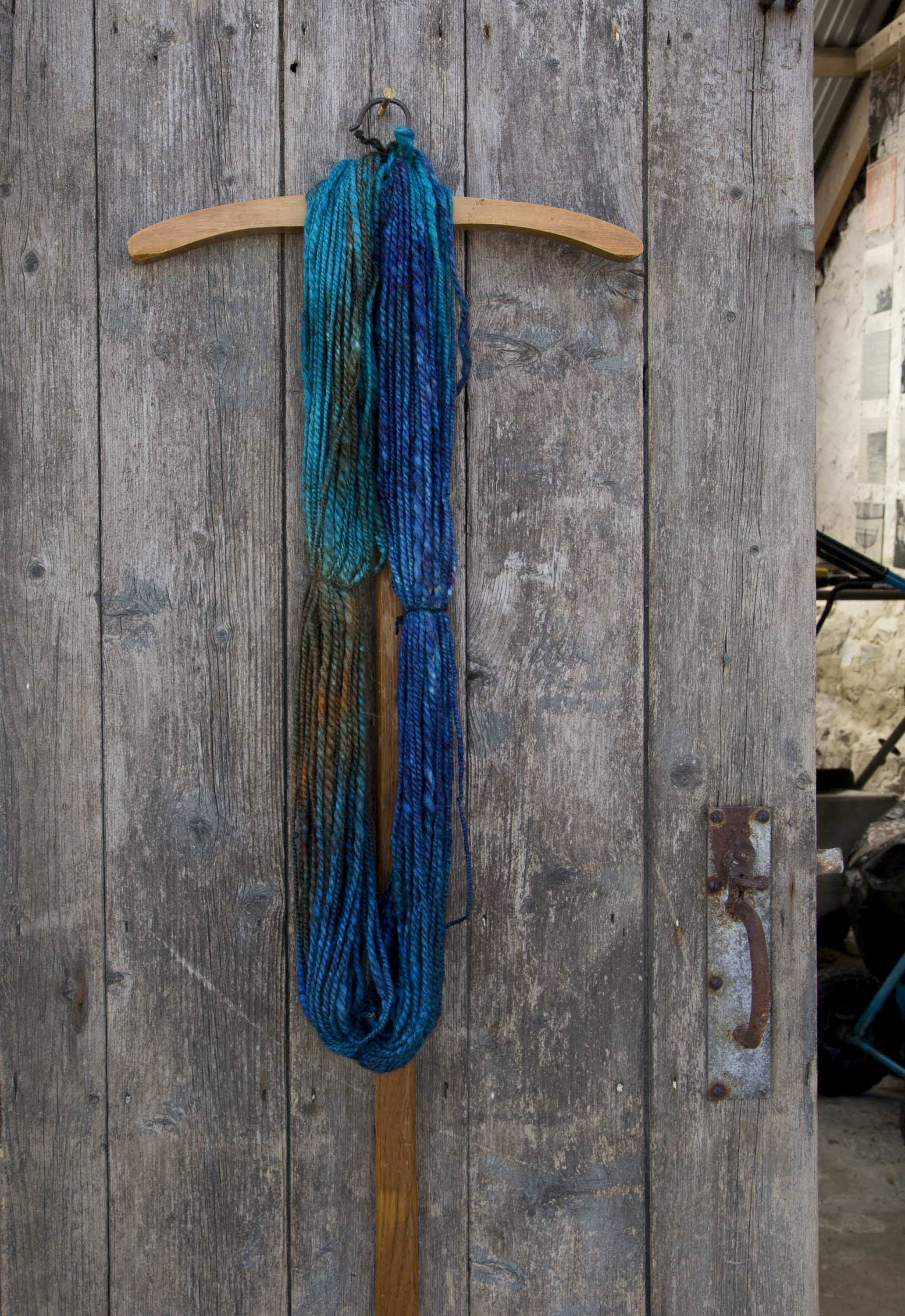 Handspun, hand-dyed art yarn made in Shetland by Niela Nell Kalra. A hank of yarn hangs on a weathered wooden door, with a glimpse inside a stone built traditional Shetland croft house.