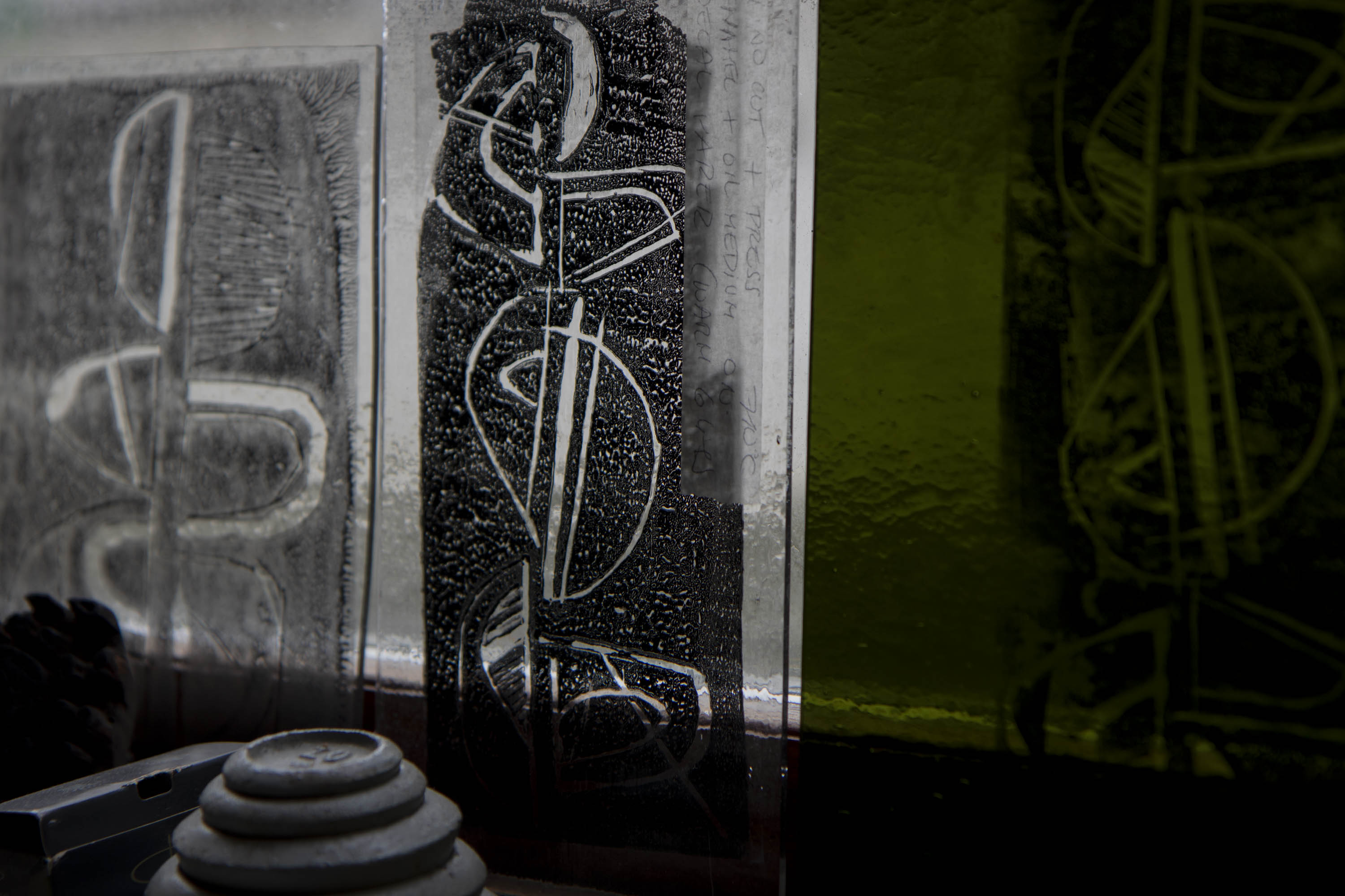 Three linocut prints on glass against a window. The ink is black and shows a reversed-out curvilinear motif. One piece of glass is limey yellow-green