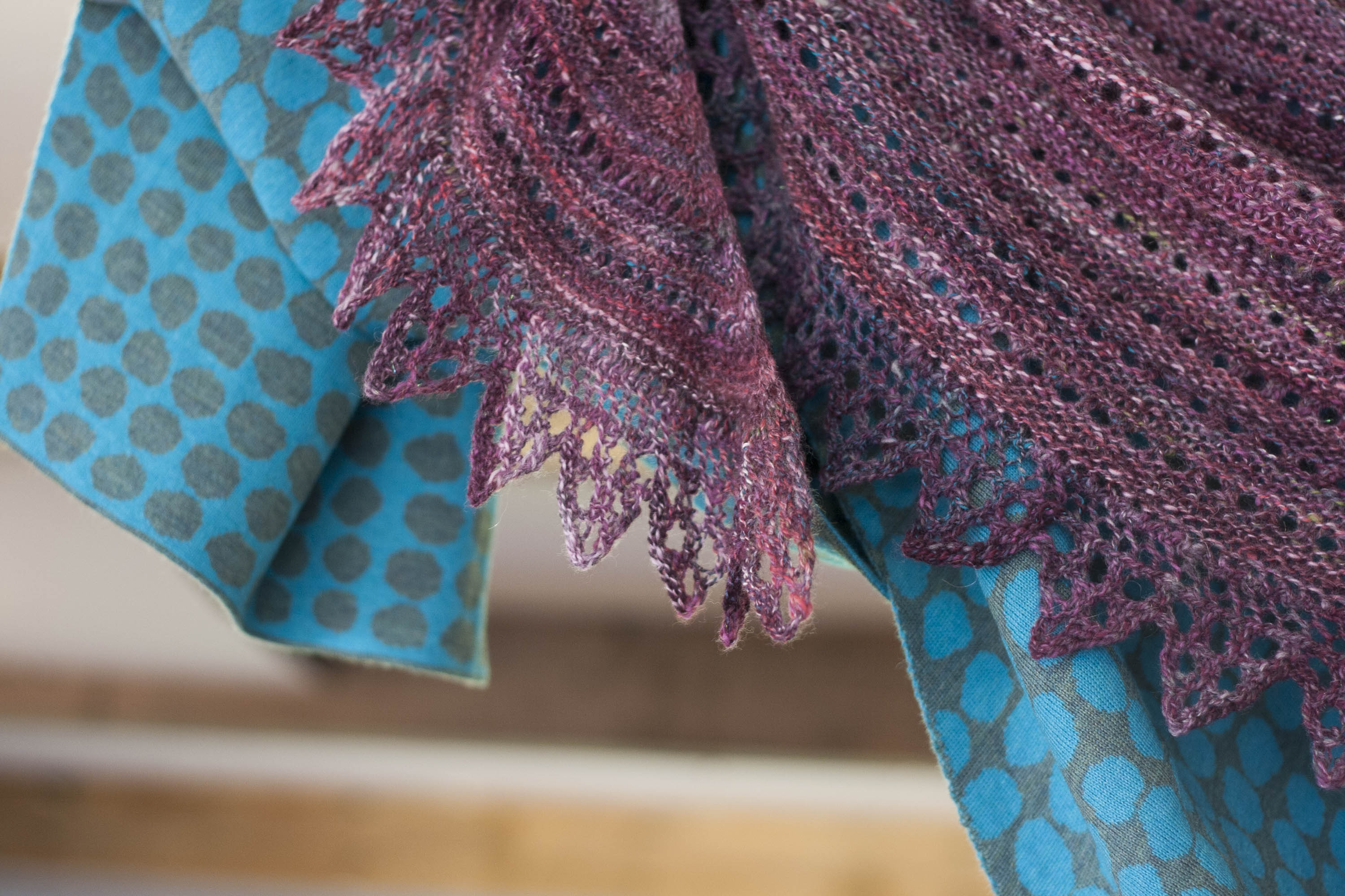 Handknitted shawl in handspun yarn in purples and plums. With lace edging. Hangs over a contemporary Nielanell shawl in blues with an abstract pattern.