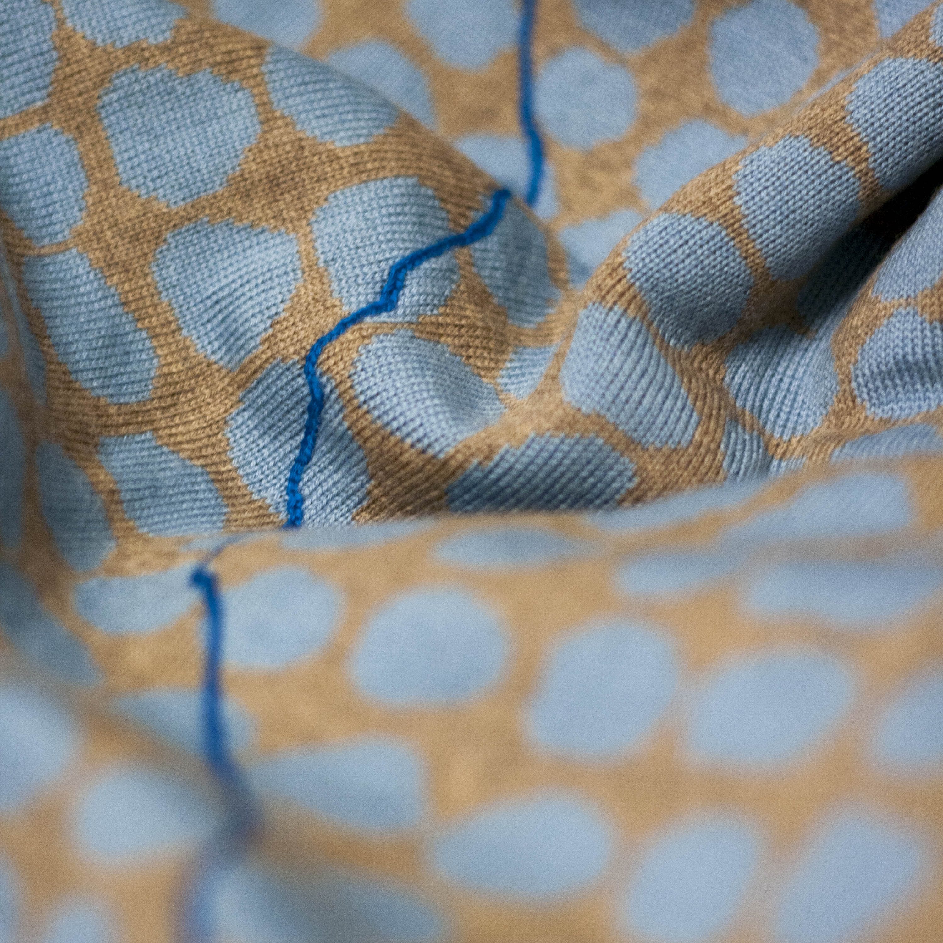 Detail of contemporary Scottish textile - a linked line of cobalt blue travels across an irregular pattern of pebbles in ochre and skye blue