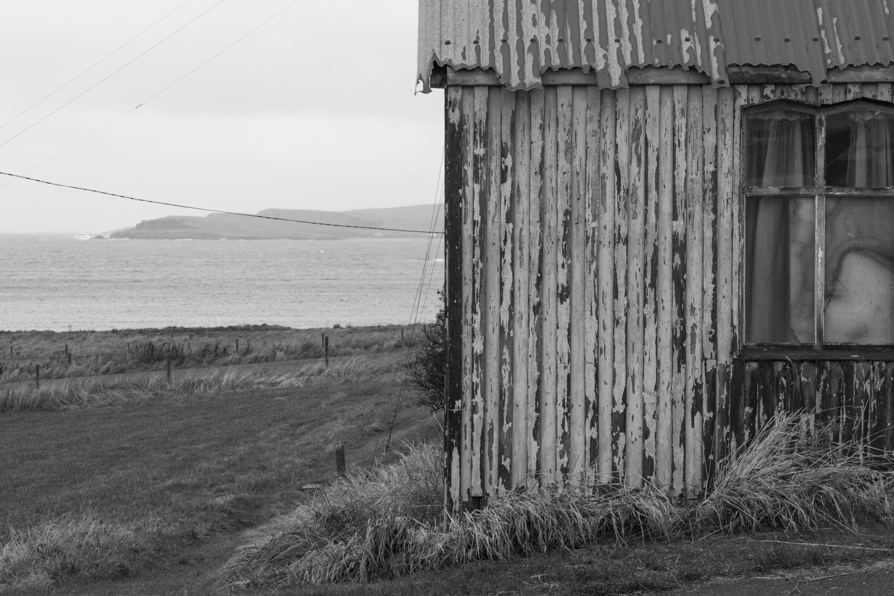 Gospel Hall in Hoswick, Shetland. A small, old wooden building with a corrugated tin roof. The tin is worn, with a jagged edge. Behind a view to the sea and Levenwick
