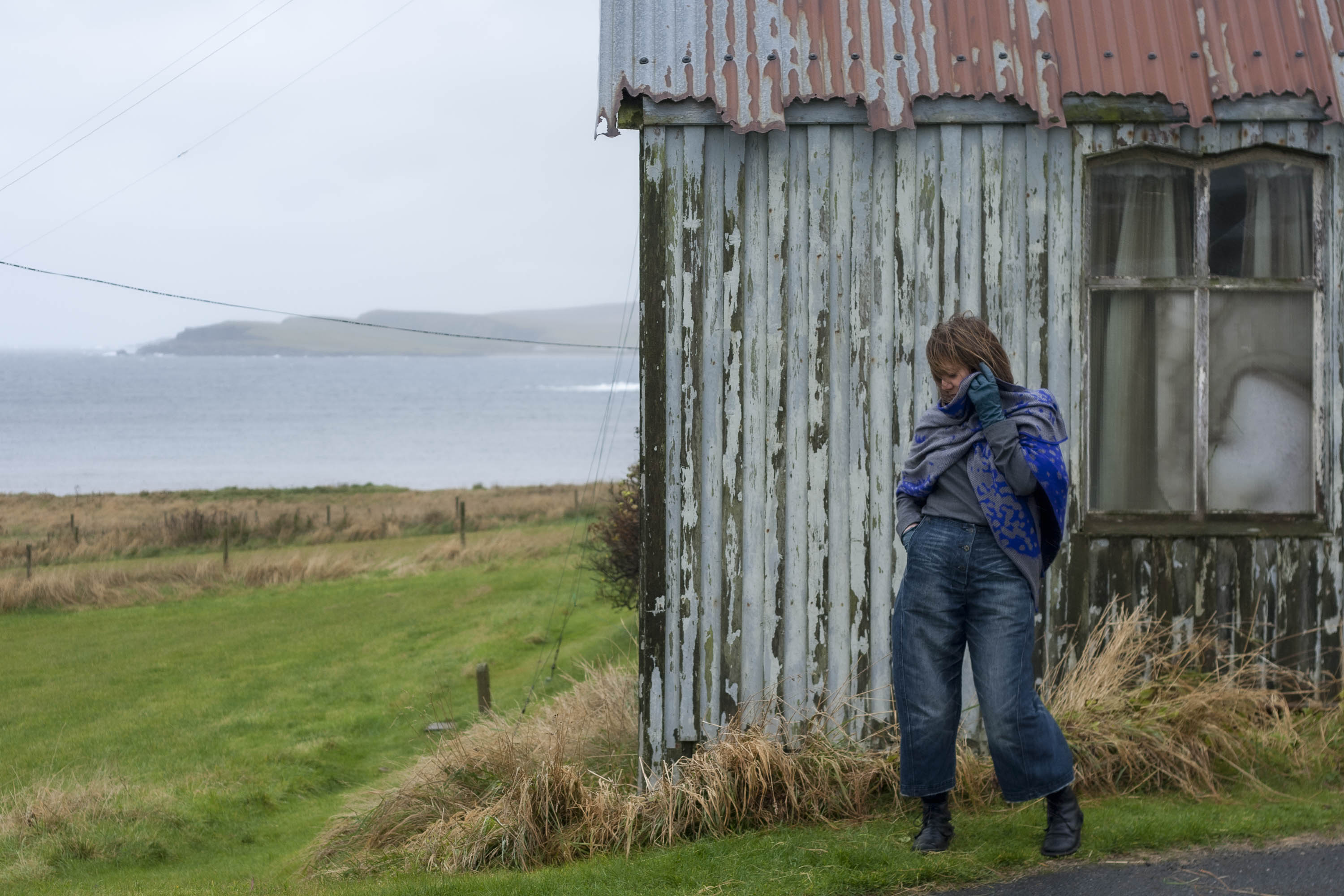 At the Gospel Hall, Hoswick, Shetland - a small, old wooden building with a rusted tin roof. In front of the building a woman stands with a grey and electric blue wrap and indigo loose jeans