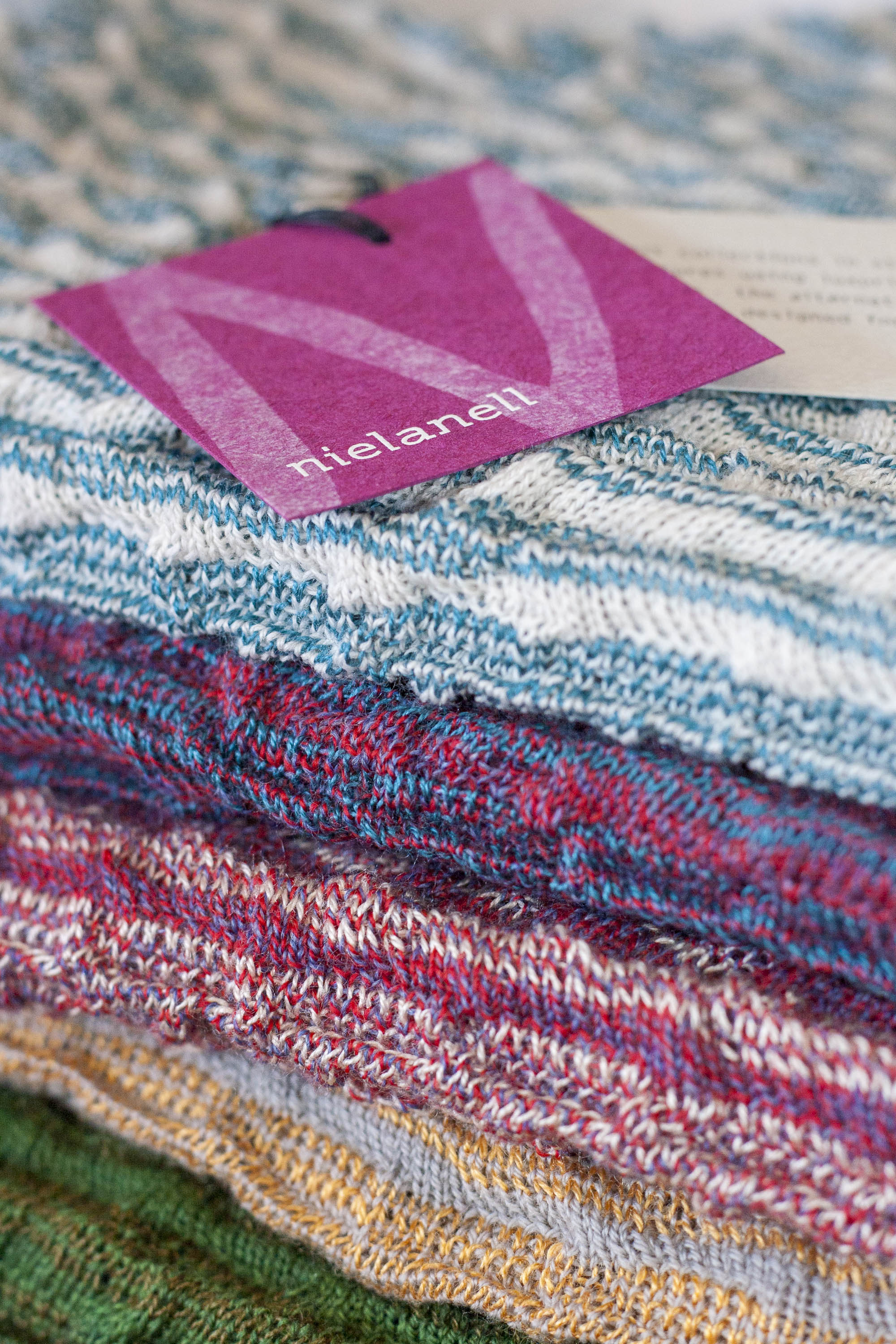 Stack of contemporary, colourful knitwear in the Nielanell studio, Hoswick, Shetland. Greens through pinks to blues. A swing ticket in recycled card is on the top garment. Pink card with white N and logo.