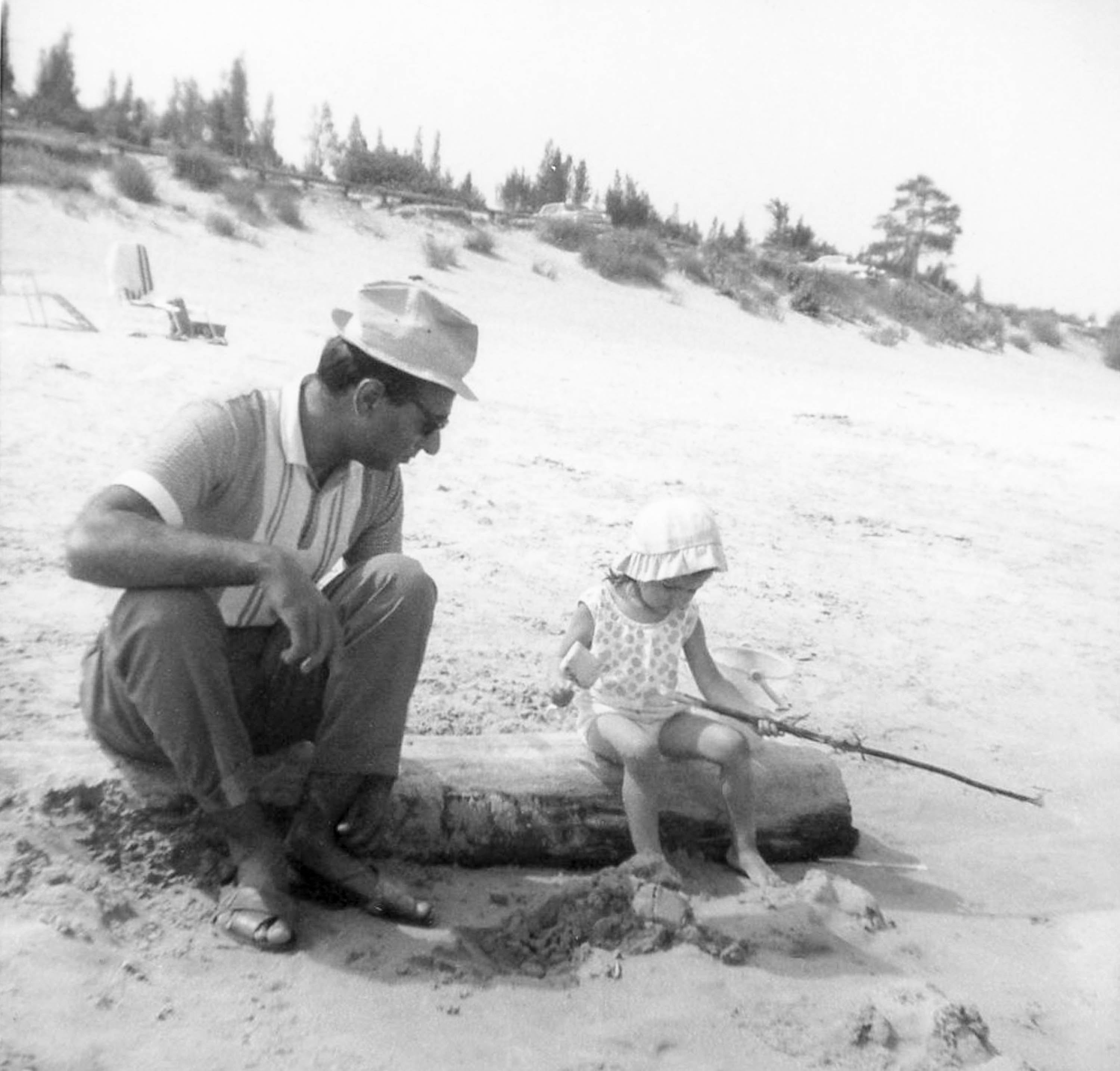 A father and his young daughter play at fishing on a beach. They sit on a log using a stick as a log and are dressed for summer, both wearing hats