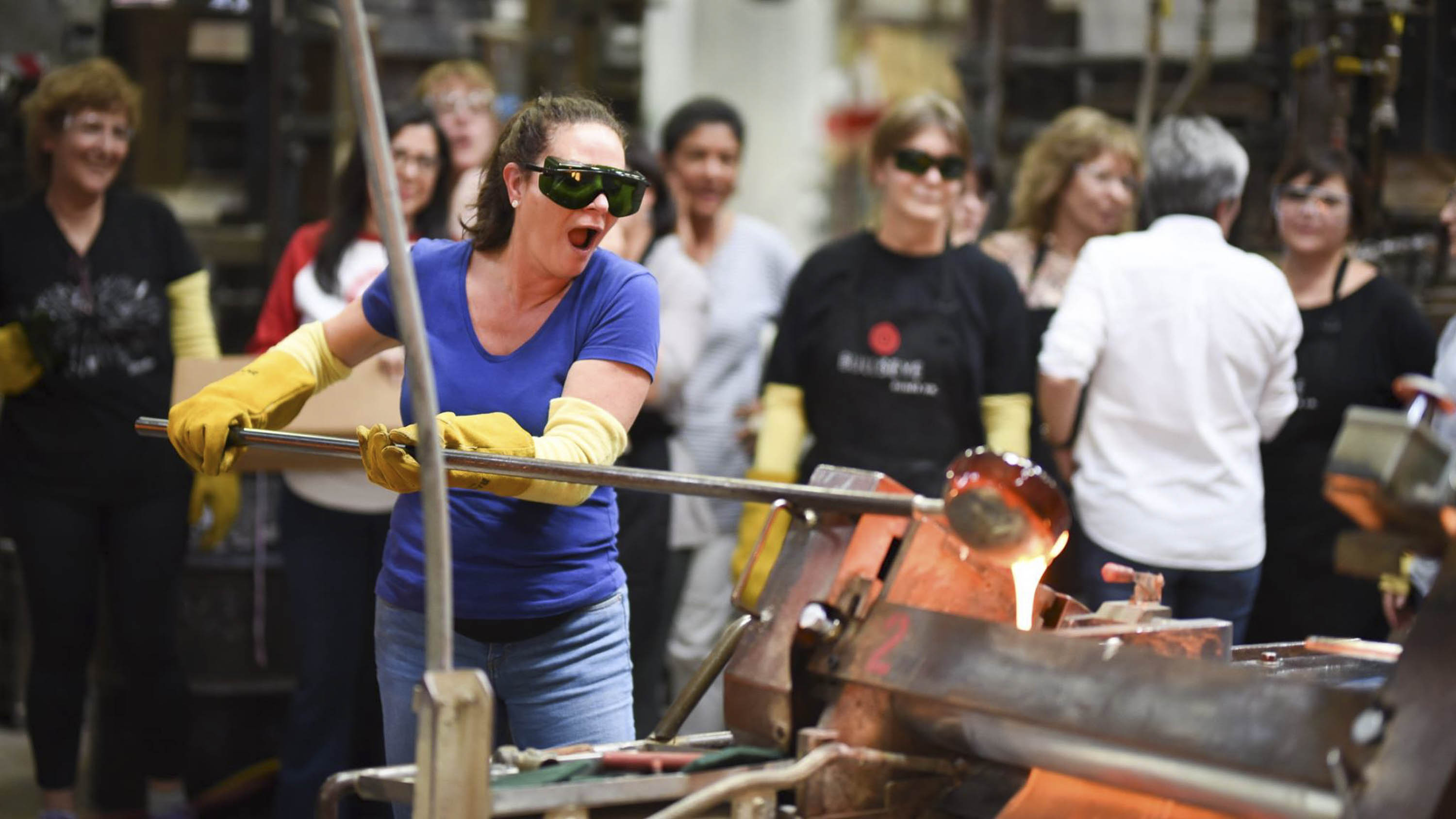 Glass-making at a workshop. A woman wearing safety glasses holds a long rod with molten glass on the end