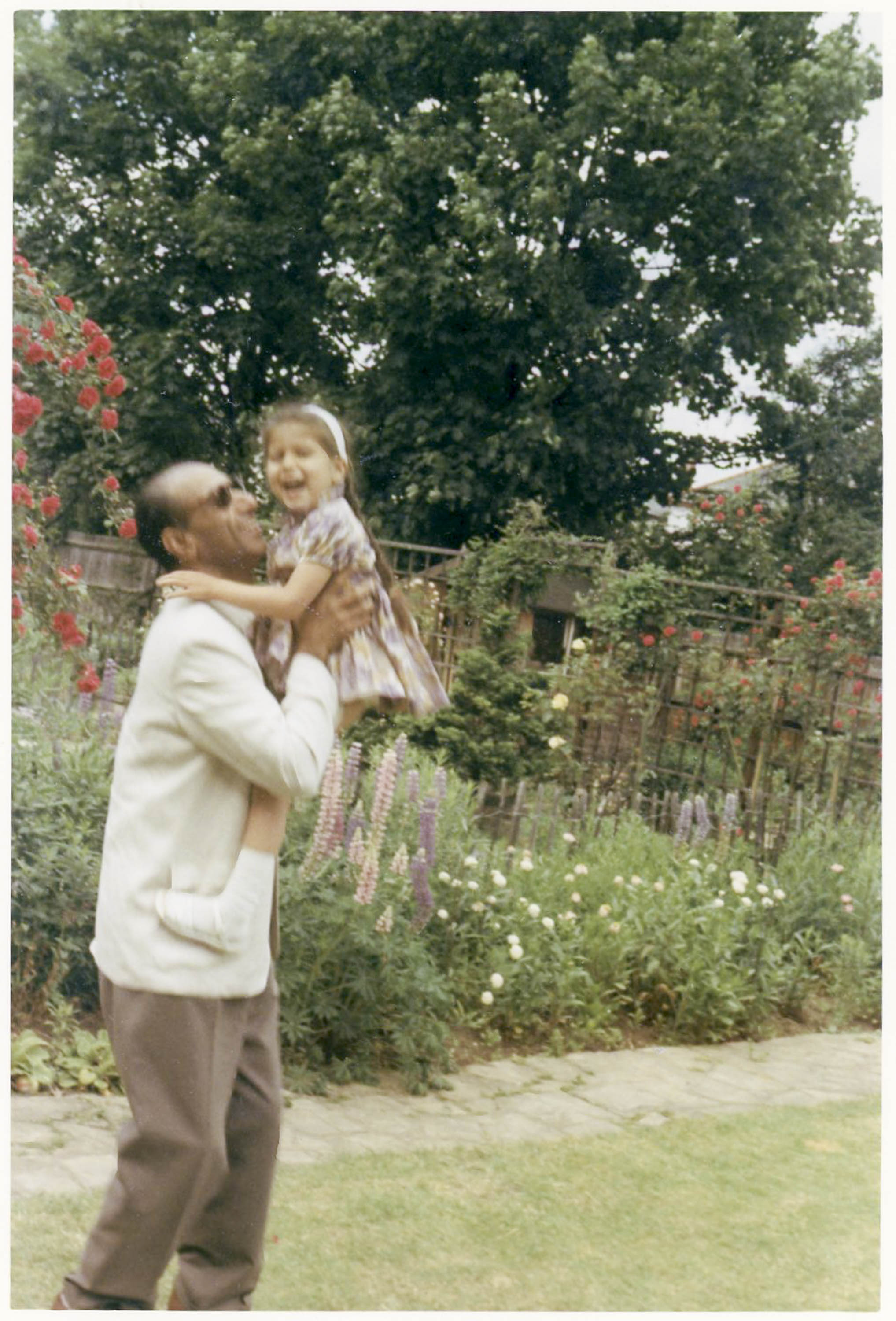 A young girl and her father play in a garden, he lifts her up high. Herbacious border in background. She is wearing a summer dress, he is in brown slacks and a cream cardigan.