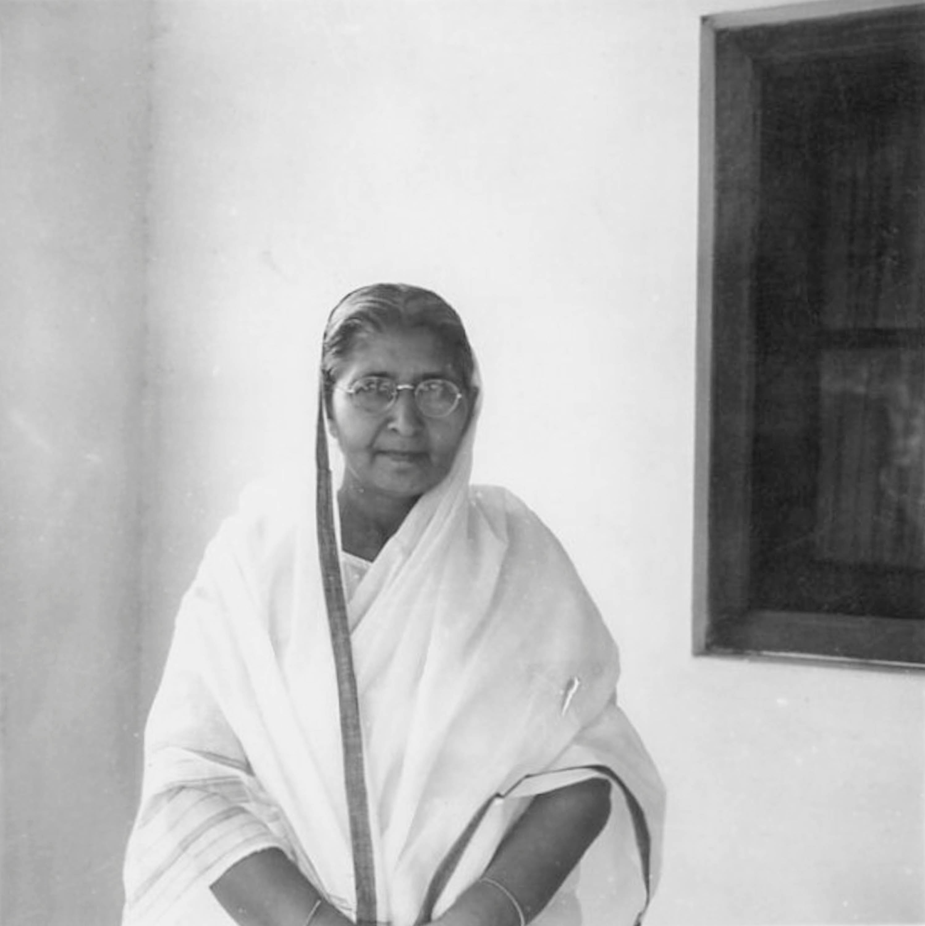 An older indian woman wearing a white sari, in front of a whitewashed wall and window