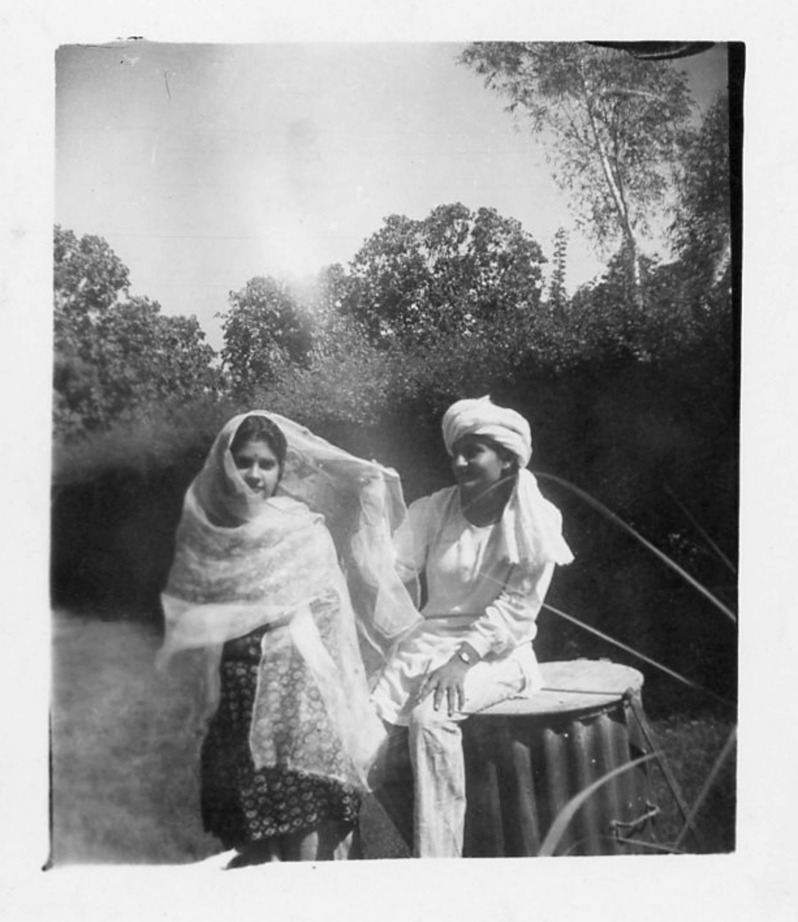 Black and white image of a young man and woman in a garden, in India. She wears a fine shawl around her head and shoulders, he wears white scarf around his head