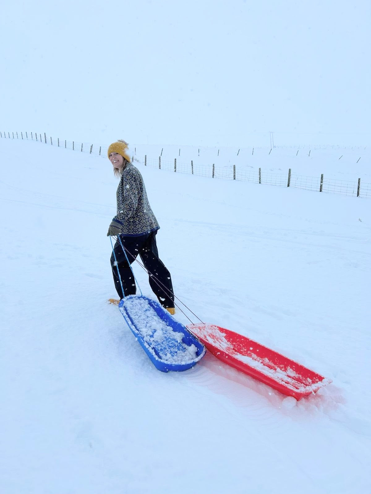 A white woman pulls a sledge up a snow covered slope in Hoswick, Shetland. She is wearing an allover Fair Isle jumper in yellow and dark petrol blue, a yellow bobble hat, black waterproof trousers and yellow wellies.