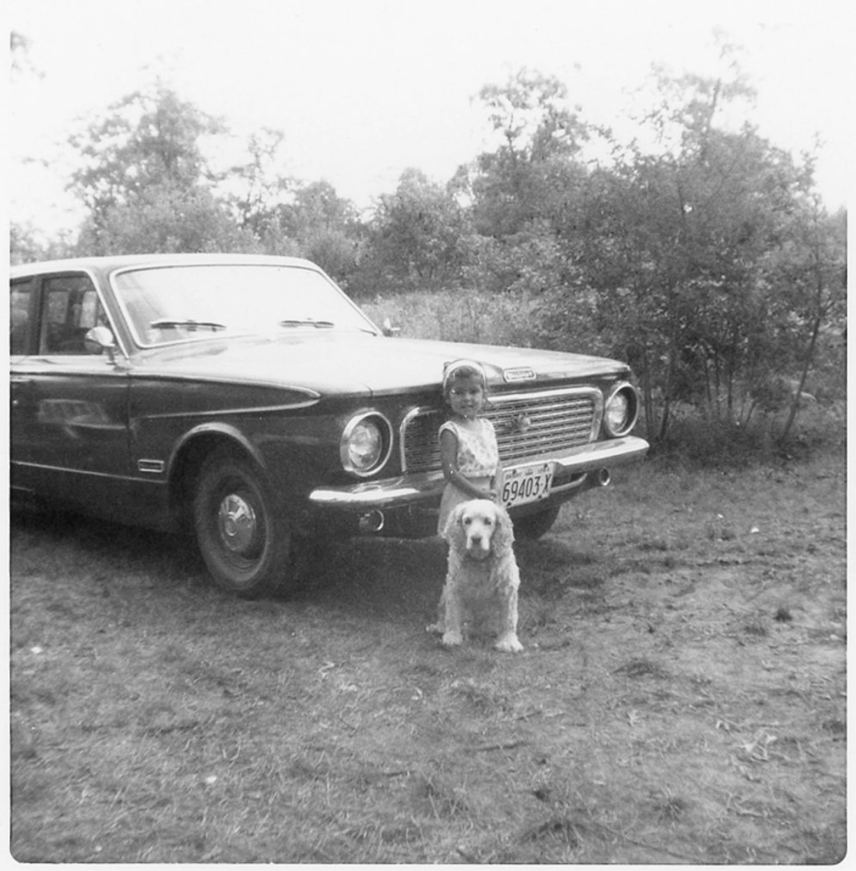Black and white photograph of young Niela, standing in front of a car with her dog - a spaniel
