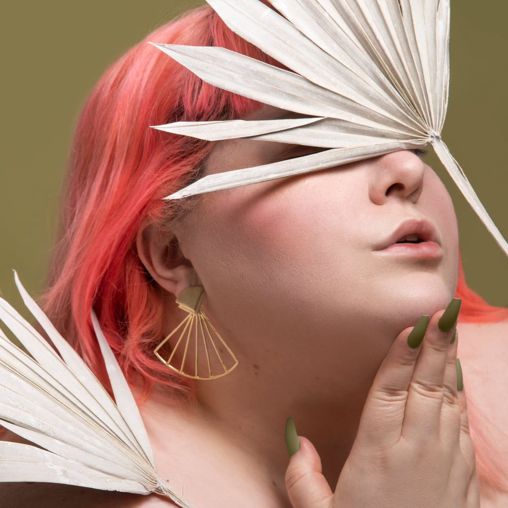 Editorial style picture of Amy Lynn, a female with pink hair. Amy's face is covered by a fan palm and she wears gold earrings and has long, olive green nails.