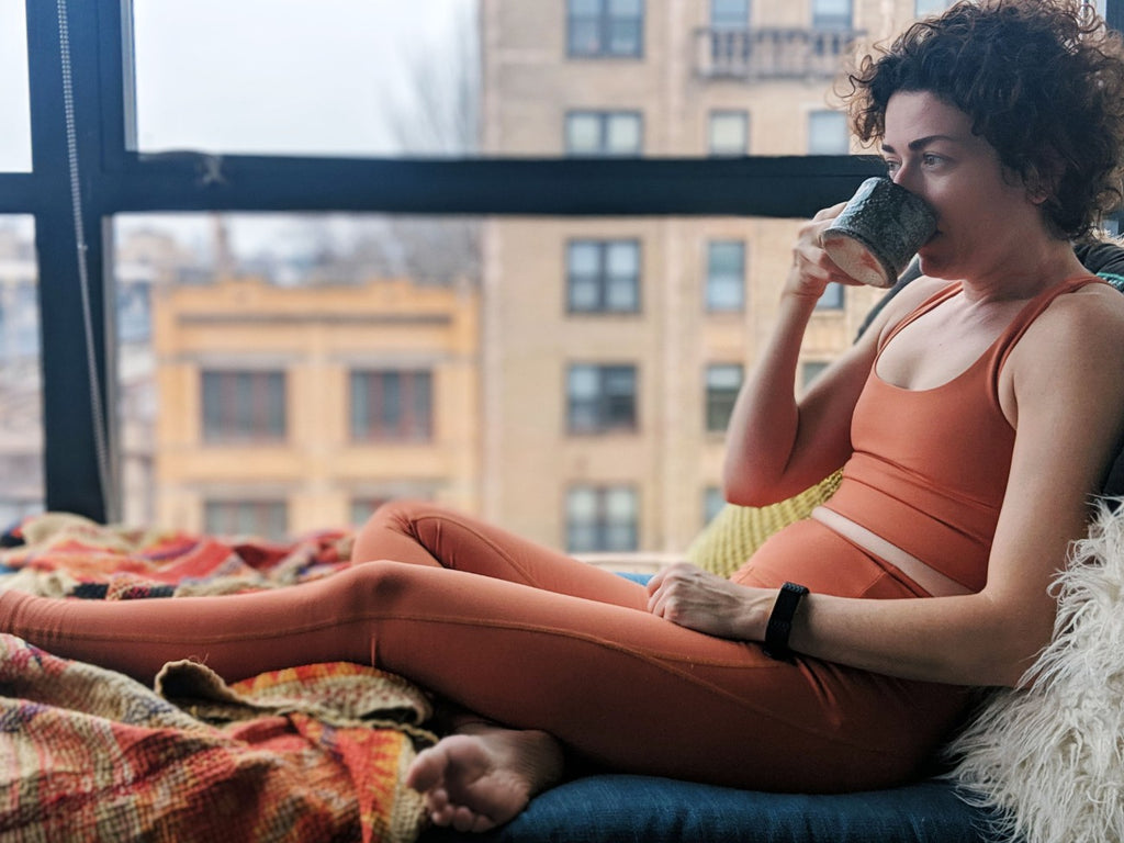 Adrianne sits on a chair, sipping coffee while wearing a workout outfit