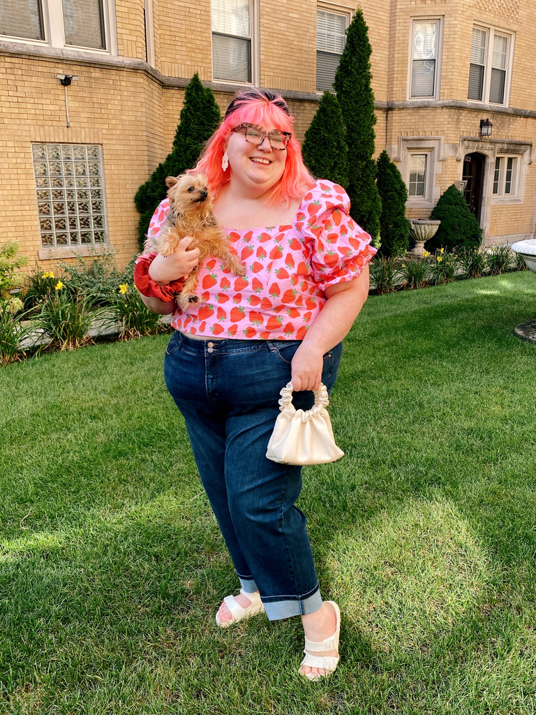 A picture of Amy Lynn, a plus sized female with pink hair. Amy wears blue jeans, a pink shirt and glasses. She holds her small dog, Stormi, in one hand.