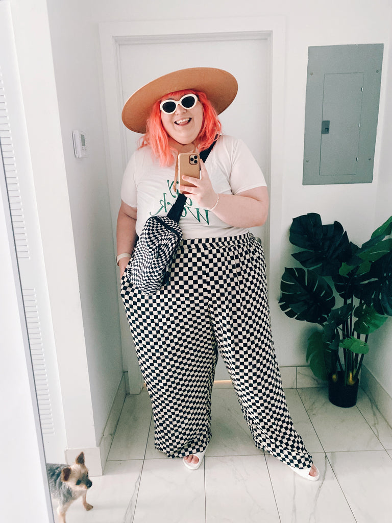 Amy Lynn, a plus sized female with pink hair, takes a selfie in a mirror. She wears round white sunglasses, a white shirt, a wide brimmed hat and black and white checked pants.