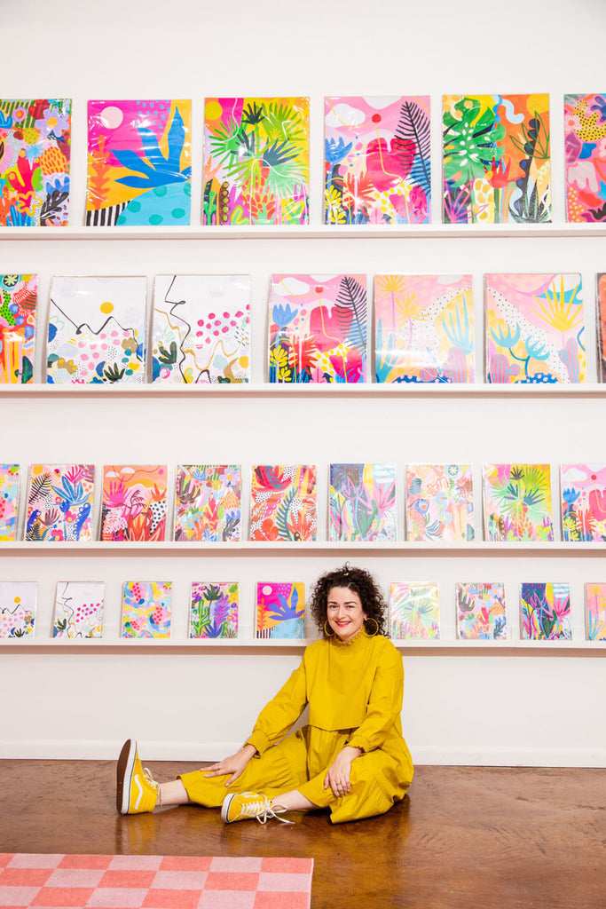 The artist sits on the floor in front of a wall of her art prints.