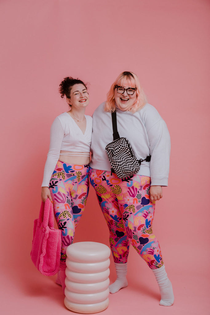 A picture of Adrianne, a white, straight sized female and Amy, a white, plus sized female with pink hair. Both wear bring pink leggings and are smiling at the camera.