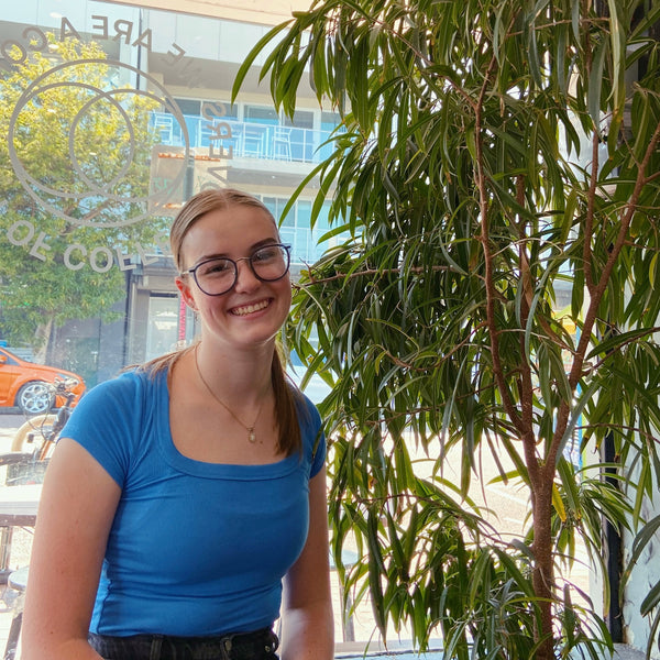 New member of the team, Alice is wearing a blue shirt and is sitting infront of a large plant in the cafe.
