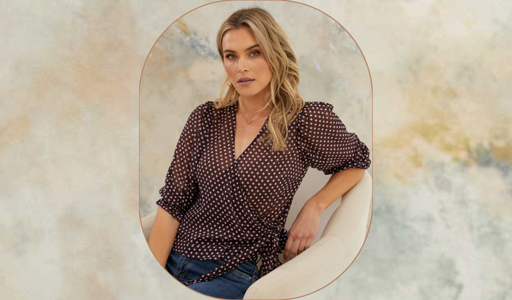 Pictured: A blonde female model is sitting on a chair, she is wearing the Caribbean Dot Top by Stella Clothing. The top features a v neckline, 3/4 length sleeves and is in a wrap style with aubergine with nude polka dot fabric.