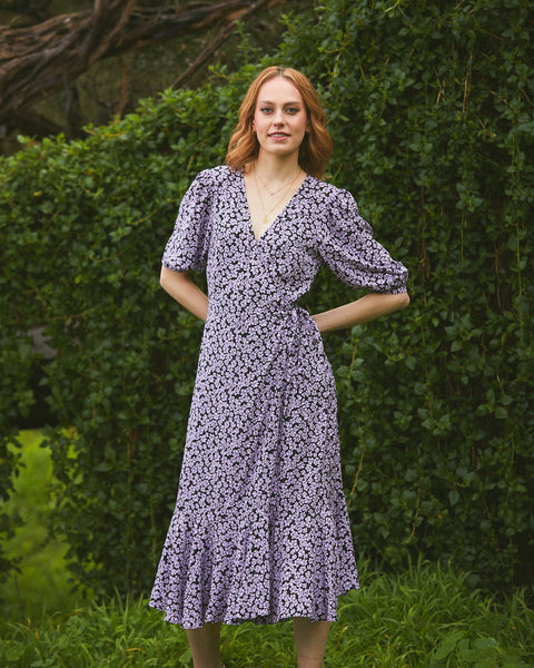 Pictured: Woman wears the Lavender Floral Dress by Stella. The dress is a wrap midi and features a v neckline.