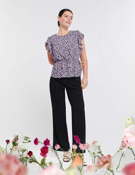 Pictured: Model is posed looking out of frame. She is wearing black pants and the Lavender Floral Top by Stella Clothing. The top is short sleeve and features a side tie synching waist. 