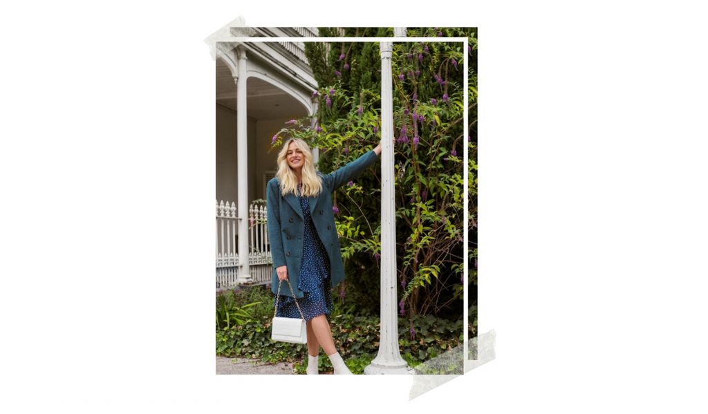 Model Brooke Mcauley is posing in front of a tree with purple flowers. She is wearing a blue dress and a teal coat with a white handbag and white boots.