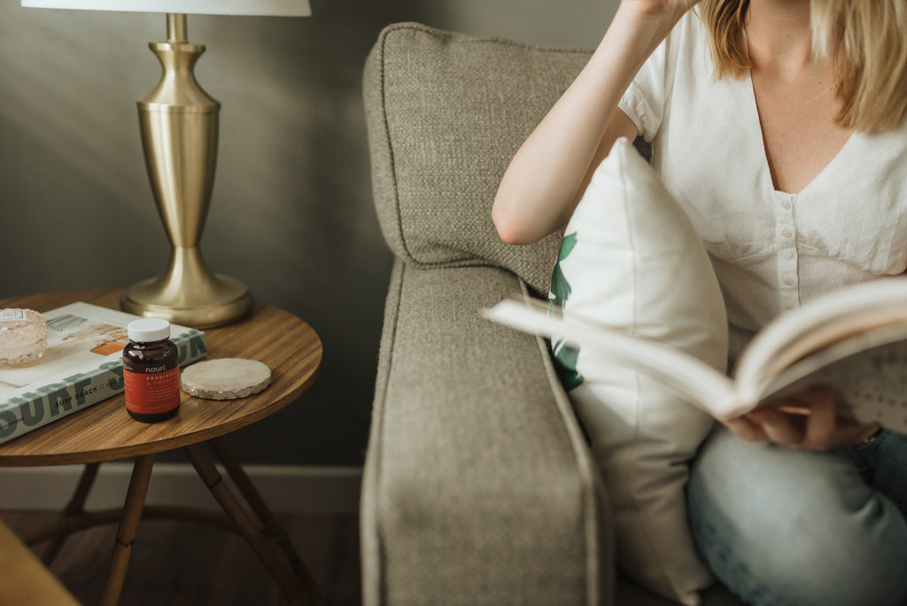 A woman sitting on a couch reading a book next to a bottle of probiotics 