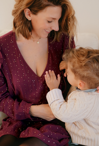 A breastfeeding mother in a burgundy Carmen nursing dress with gold polka dots in cotton gauze, responsible, made in Europe with her baby who is nursing and gives an incredible look at the camera
