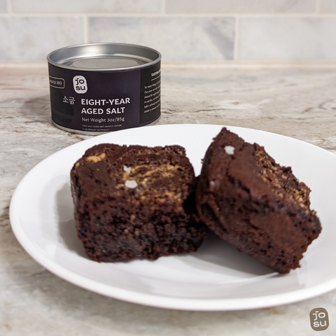 Out-of-focus close up of two Salted Tahini Swirl Brownies on a plate with in-focus jar of Josu Aged Salt in background.